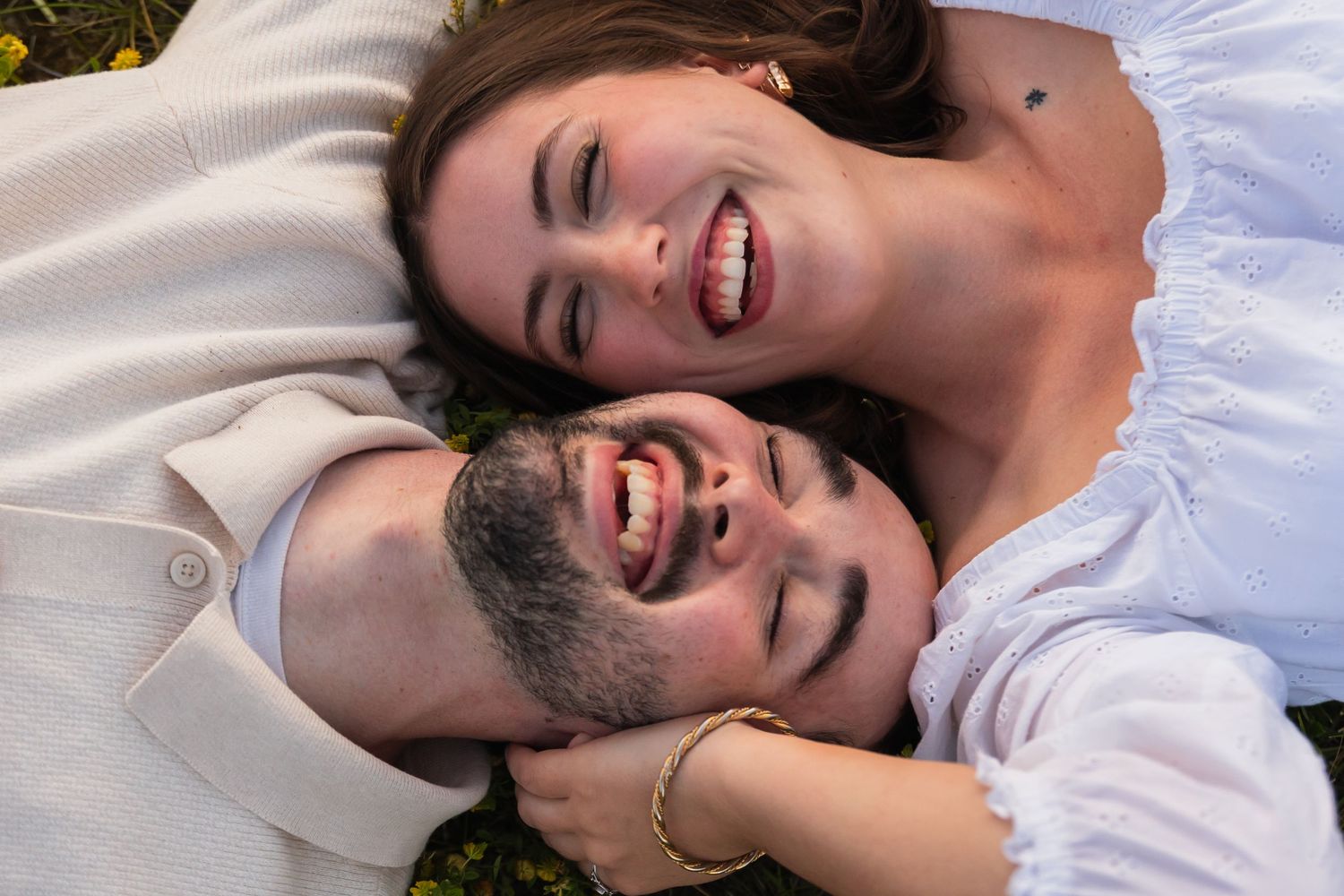 Happy couple lying on grass, smiling and laughing together, woman in white dress, man in white shirt.