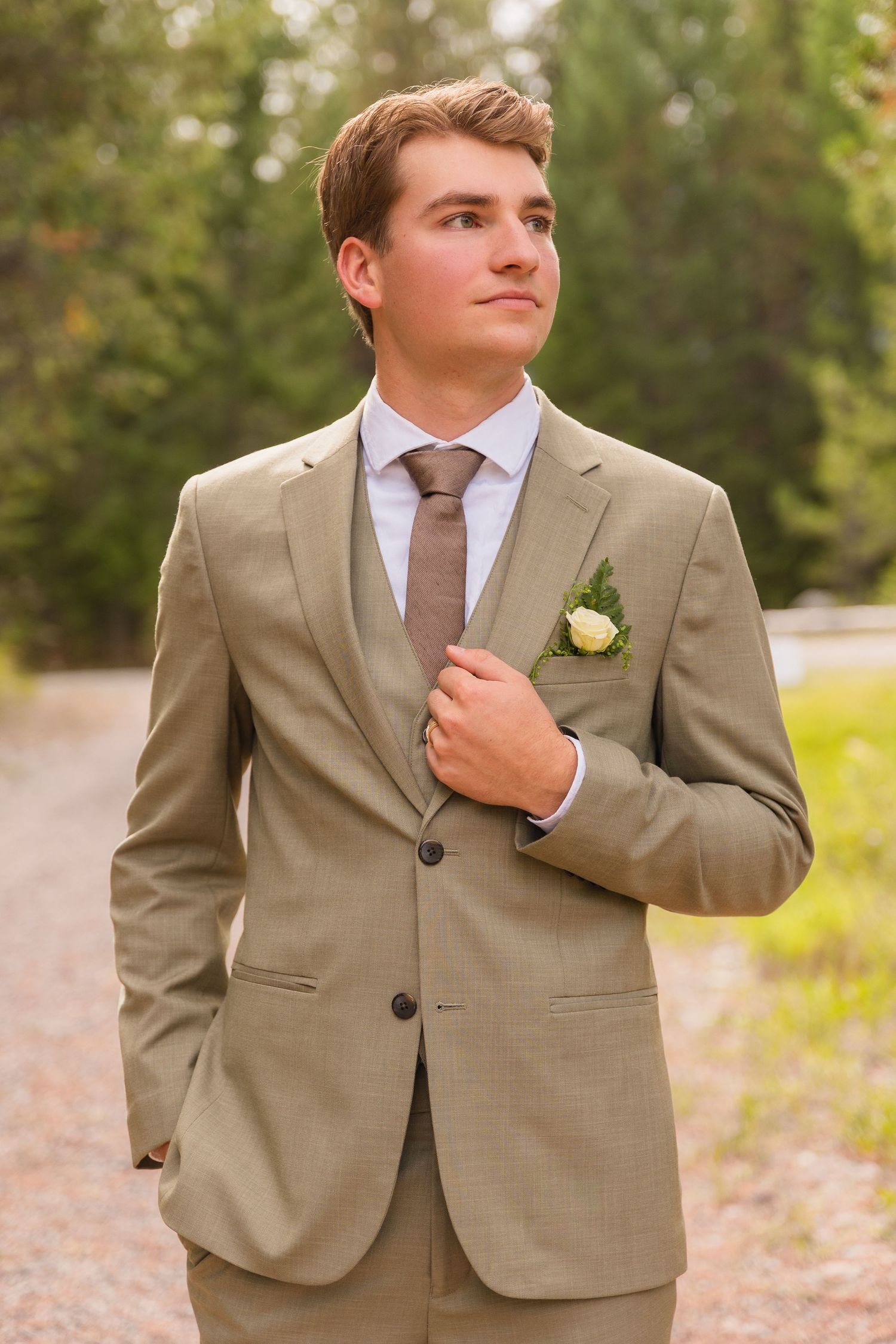 Young groom in tan suit with boutonniere standing confidently on a tree-lined outdoor path.