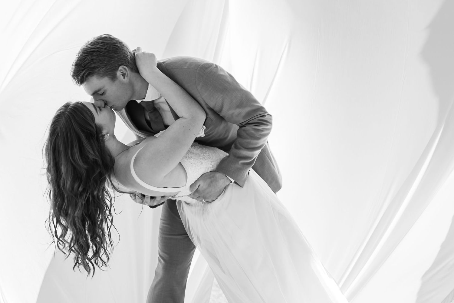 Black and white photo of a couple sharing a romantic dip kiss in a flowing white draped backdrop.