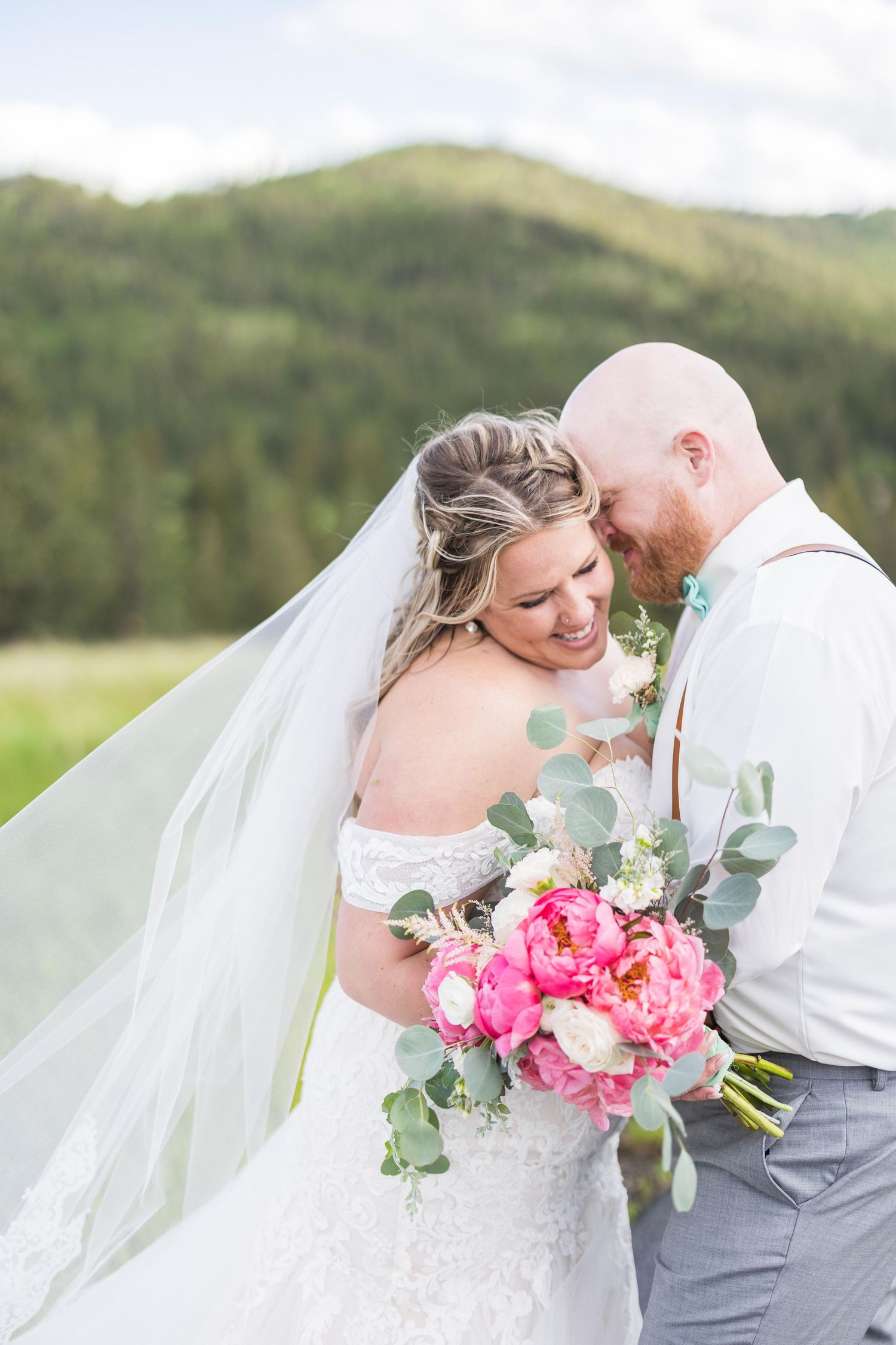 Smiling bride holding pink peony bouquet embraces bald groom with suspenders against mountain backdrop.