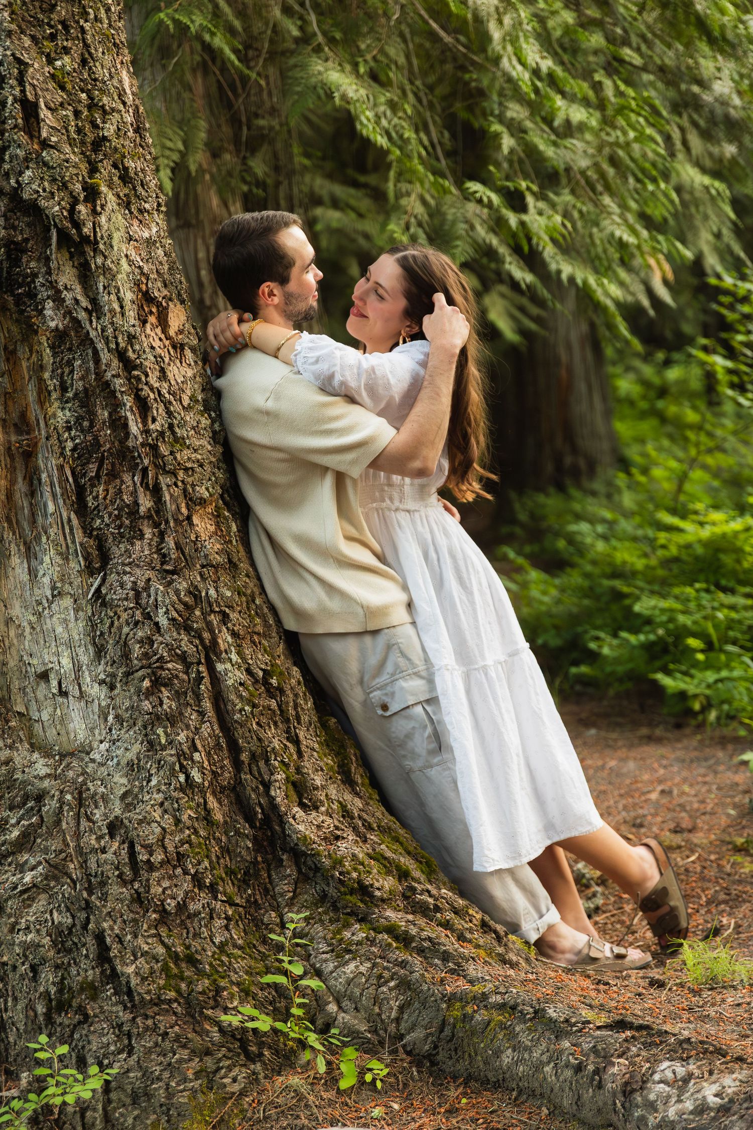 Couple sharing tender embrace leaning against large tree trunk in lush green forest.