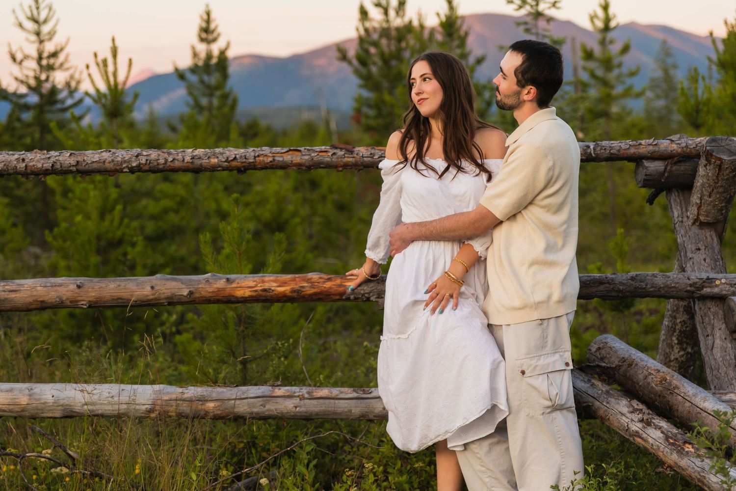 Couple embraces by a rustic wooden fence surrounded by lush green forest at sunset.