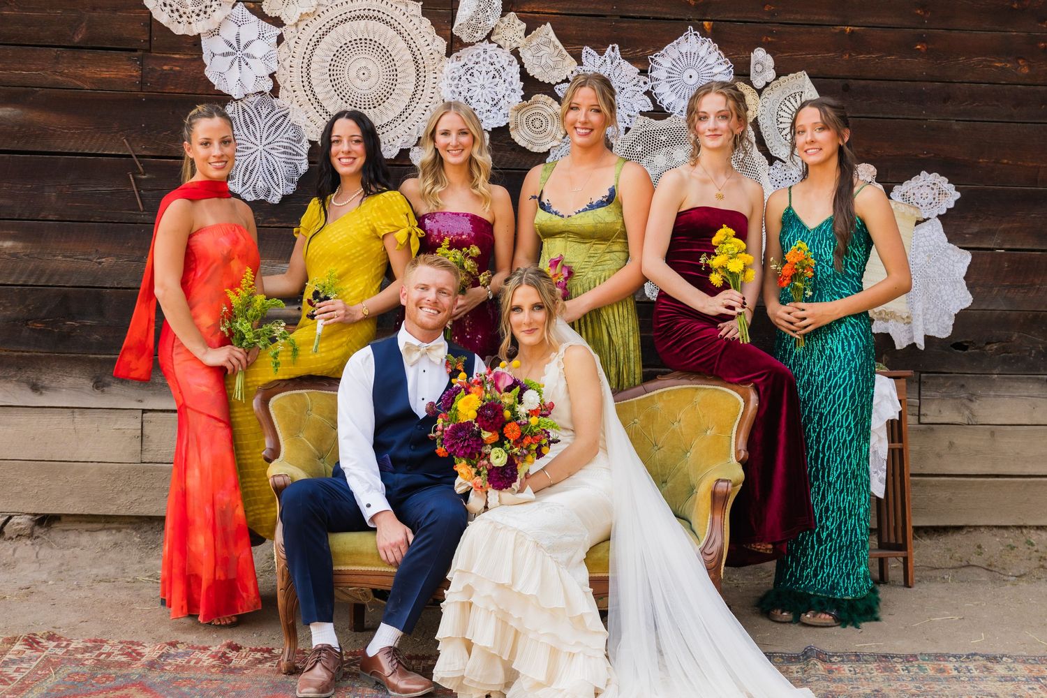 Wedding party poses with bride and groom on vintage sofa, bridesmaids in colorful mismatched gowns holding bouquets.