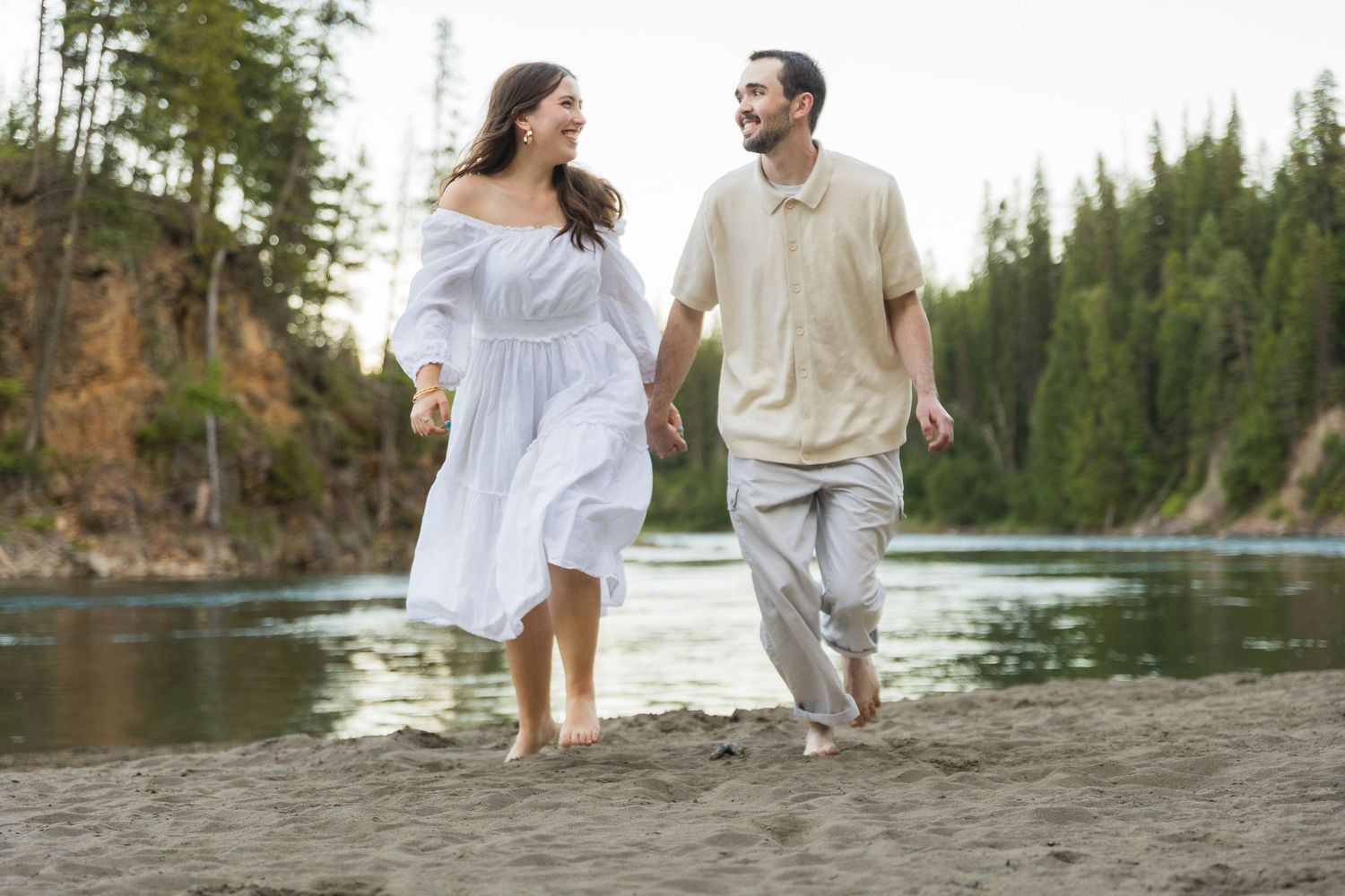 A couple walks hand-in-hand barefoot on a sandy beach, smiling at each other with evergreen trees in the background.