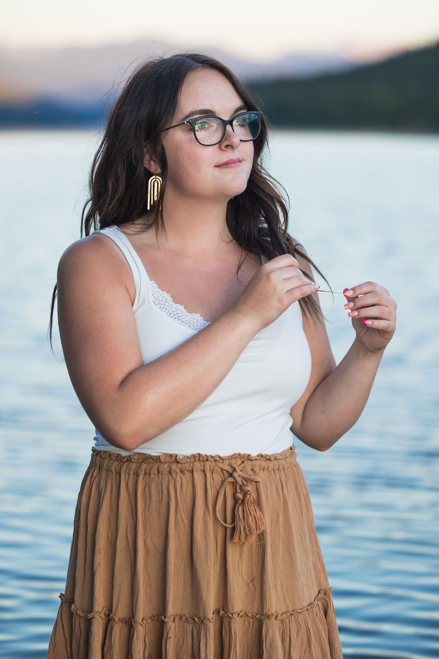 A young woman with glasses and long dark hair poses near a serene blue lake at sunset.