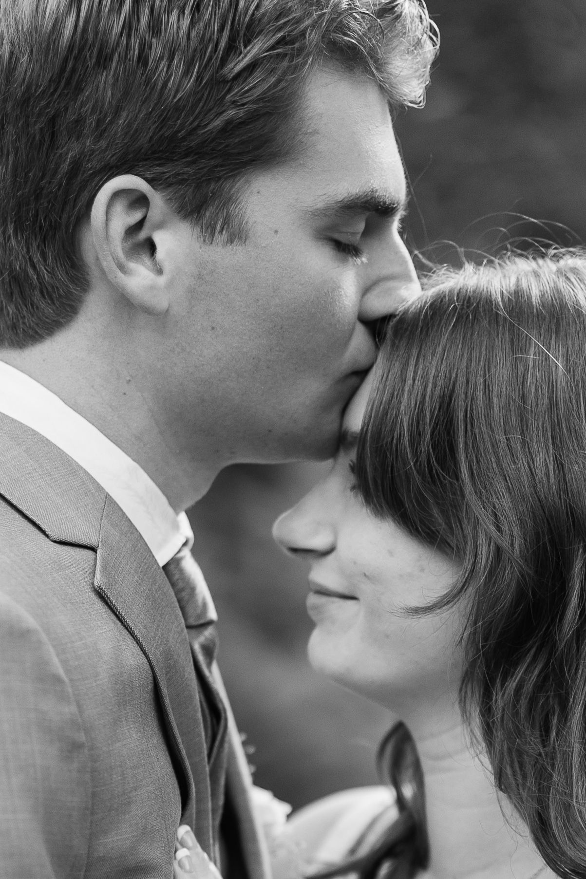 Black and white close-up of a groom kissing his smiling bride on the forehead outdoors.