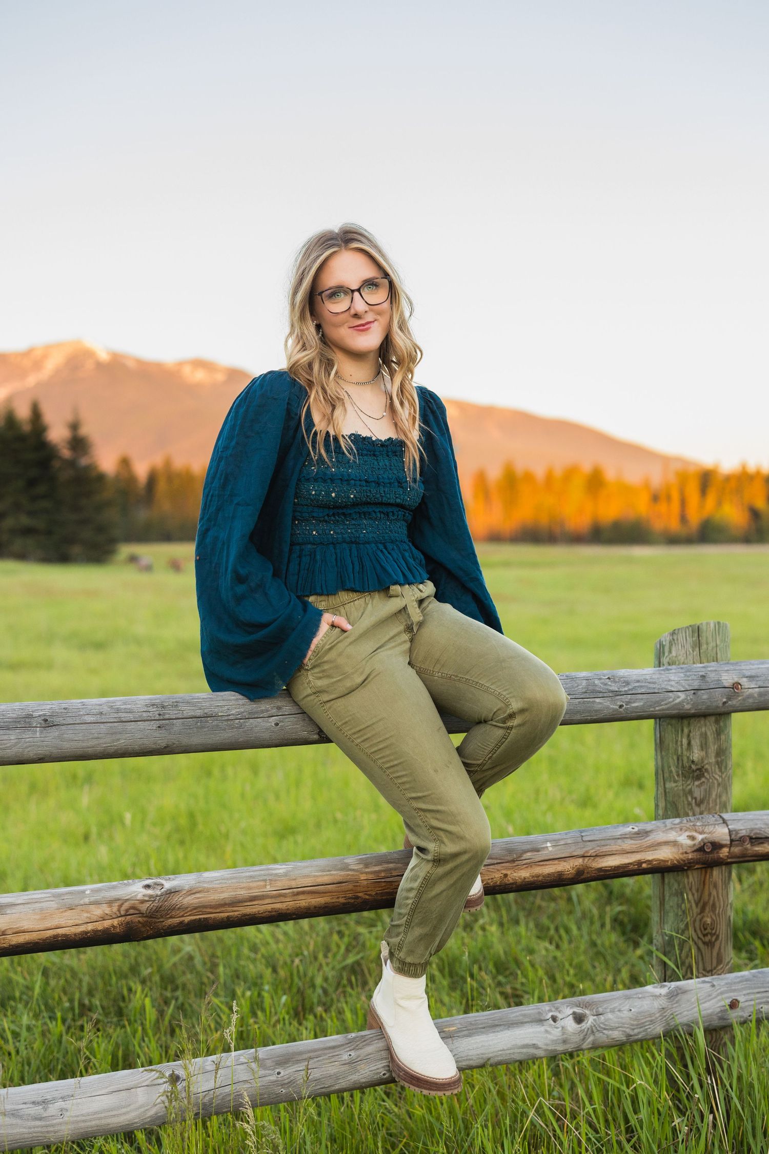 Blonde woman in teal top and olive pants sitting on a rustic wooden fence with mountain backdrop at sunset.