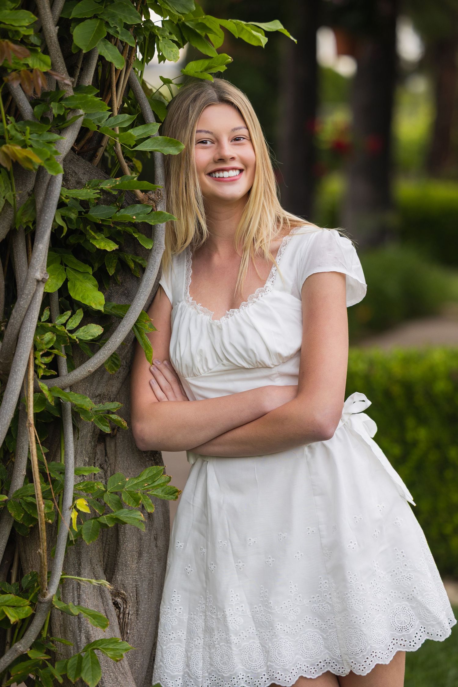Blonde woman in a white dress smiling with arms crossed beside a vine-covered tree.