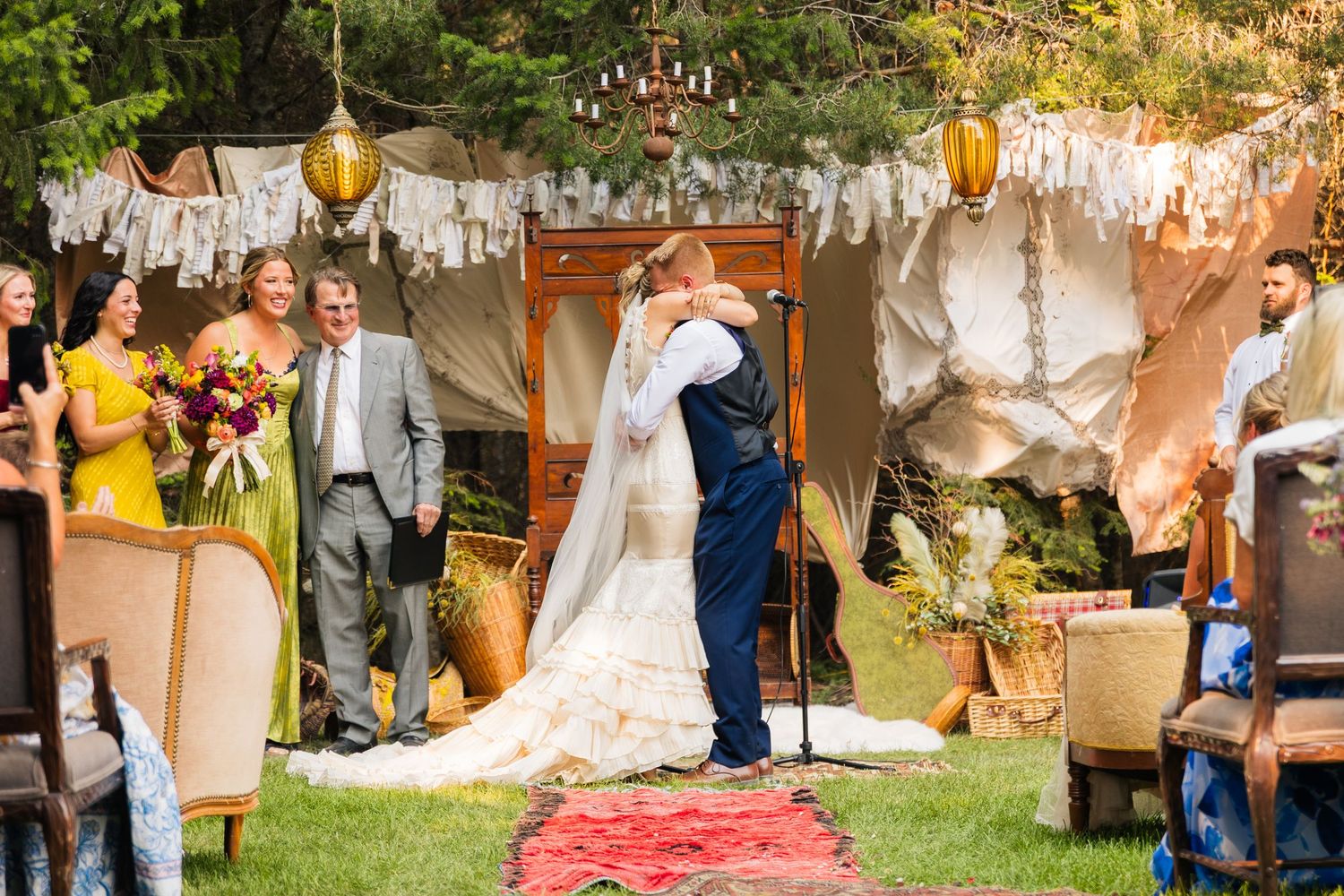 Bride and groom share first kiss at outdoor bohemian wedding ceremony with tent backdrop and golden lanterns.