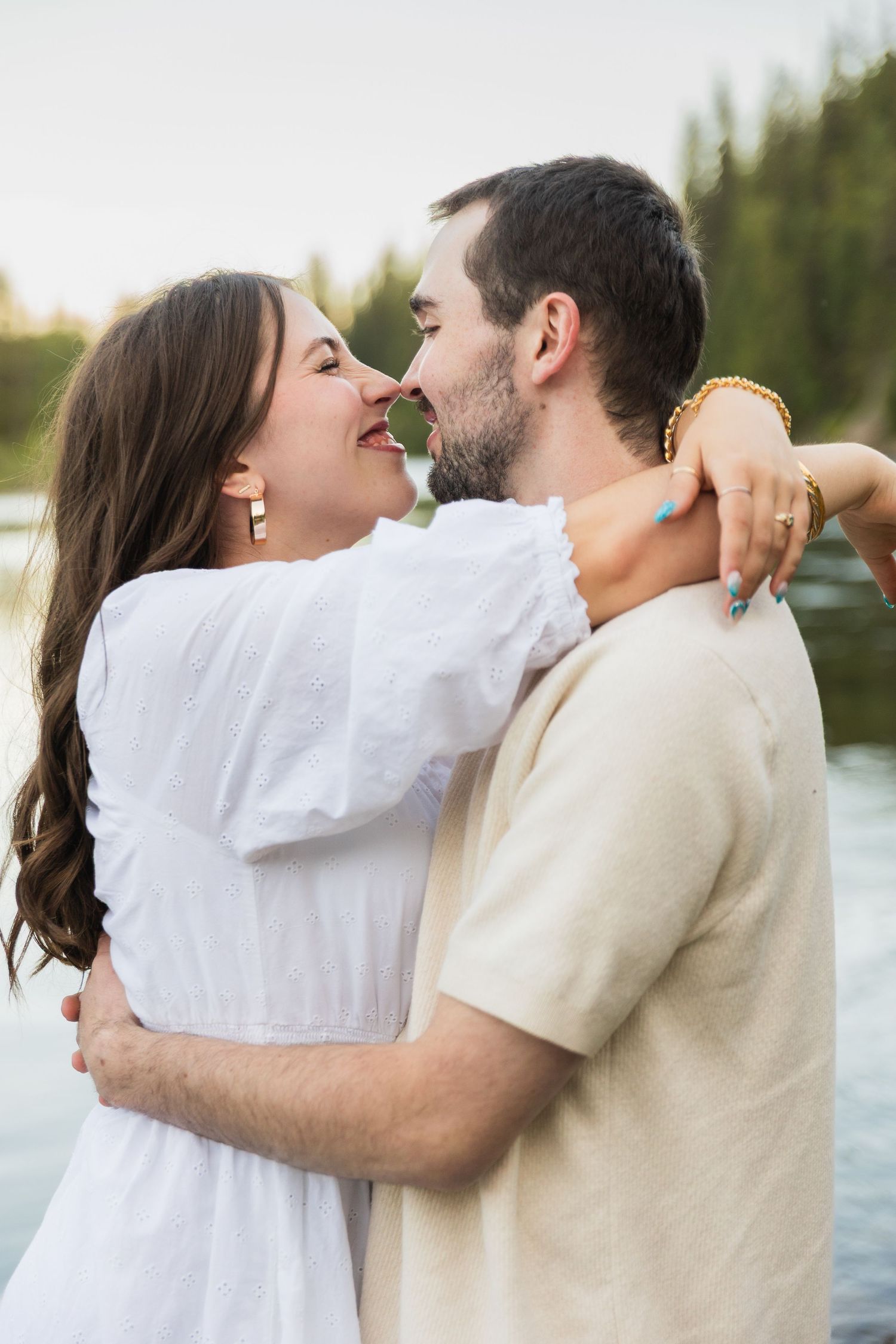 A couple embraces and touches noses near a lake, woman in white dress with arms around man in beige shirt.