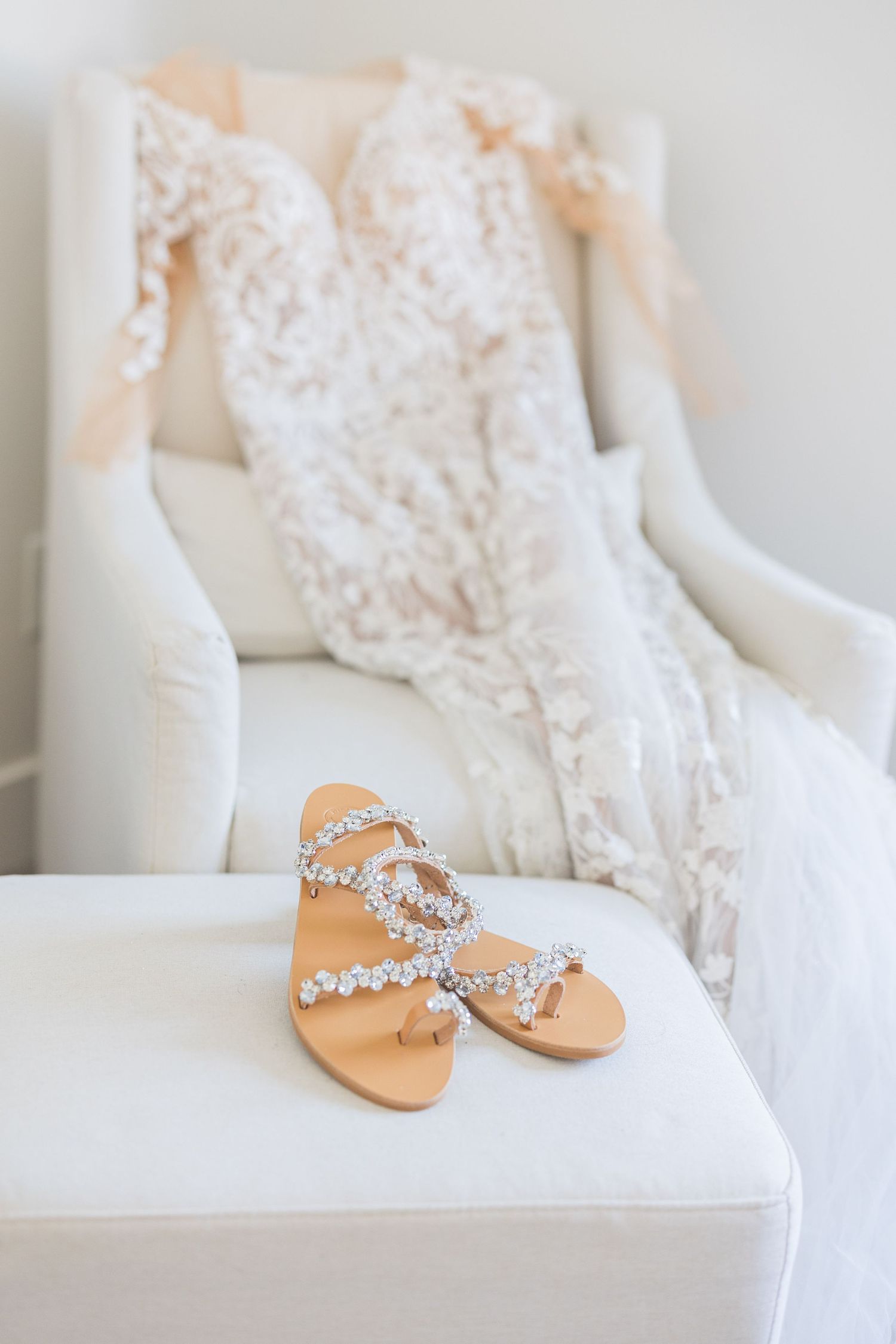 Jeweled bridal sandals displayed on white chair ottoman with lace wedding dress draped in background.