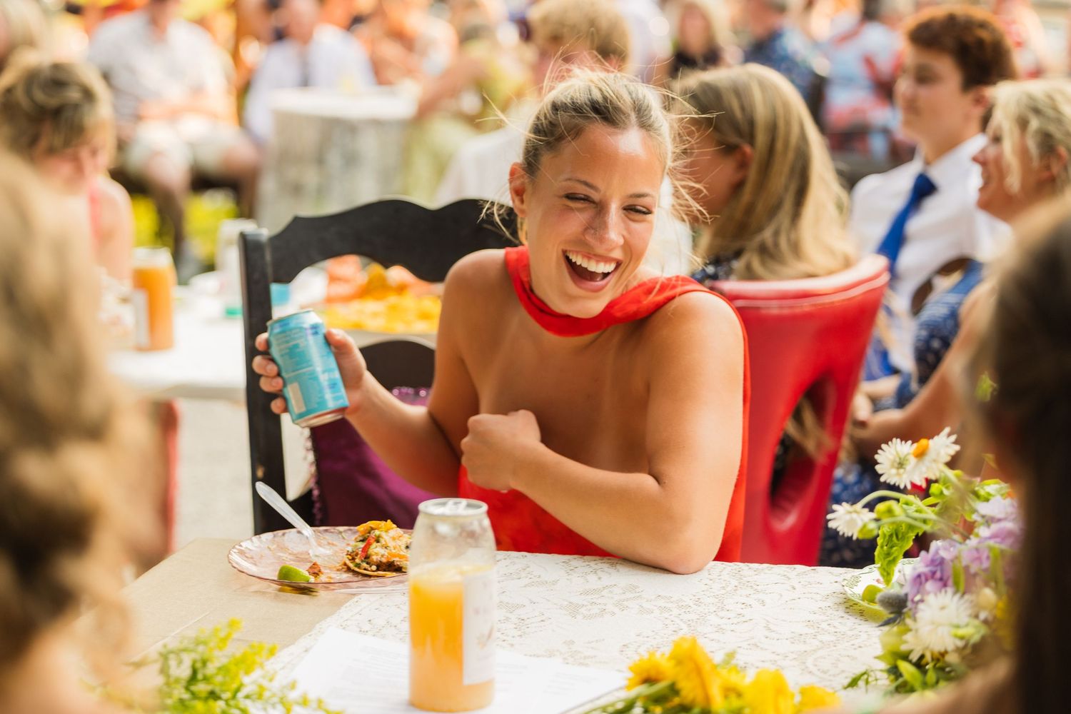 Woman in red dress laughing joyfully at outdoor party, holding teal cup at flower-decorated table.