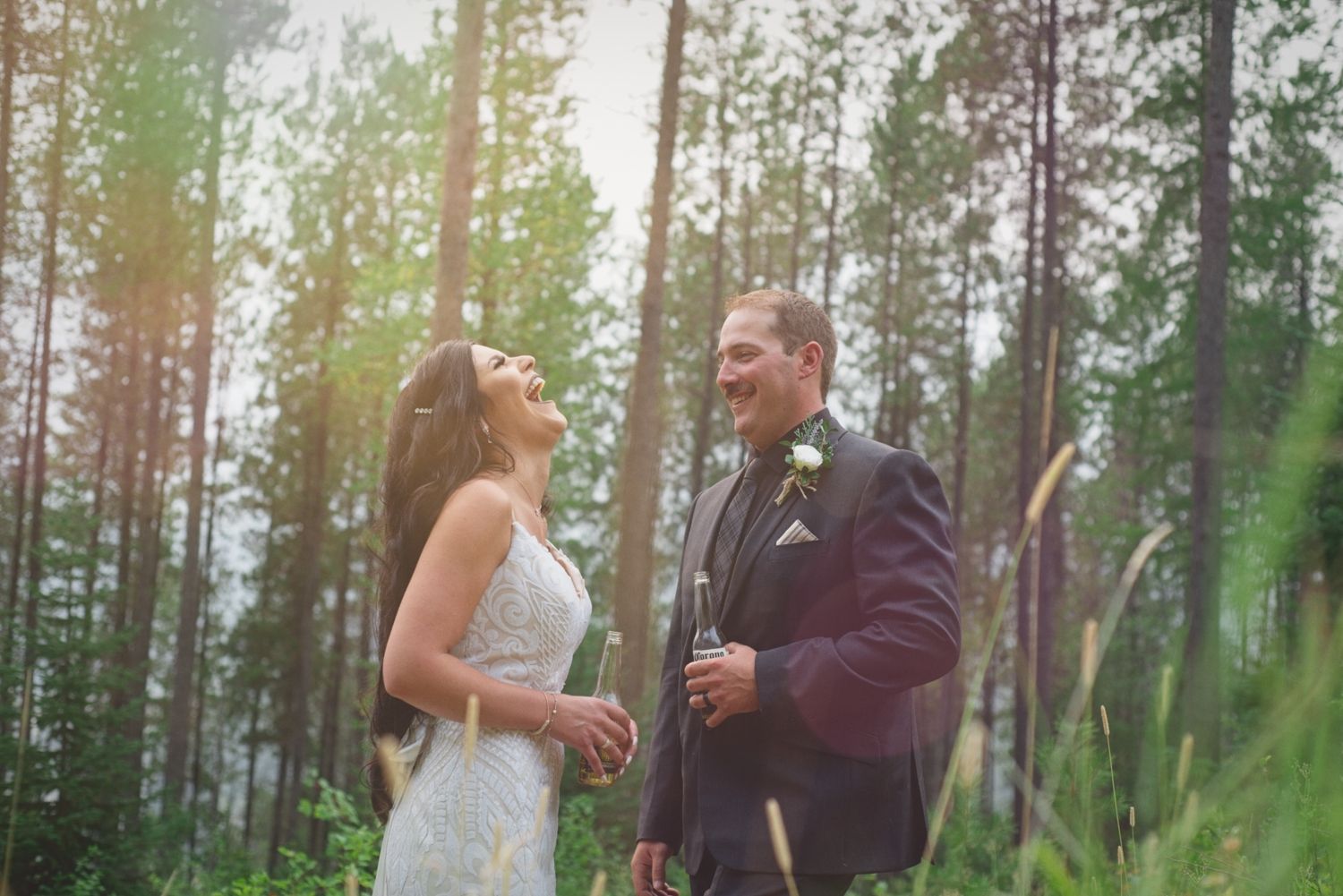 Bride and groom laugh while sharing a beer in the forest after their wedding ceremony.