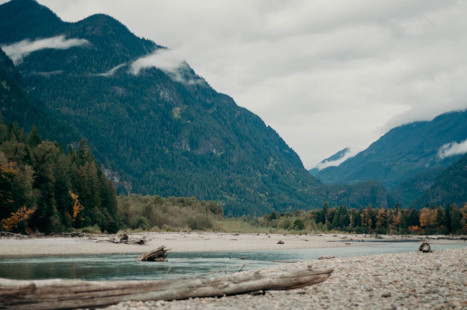 Shot of rocky beach, turquoise river, green forest, navy mountains, and cloudy sky in Squamish, BC.