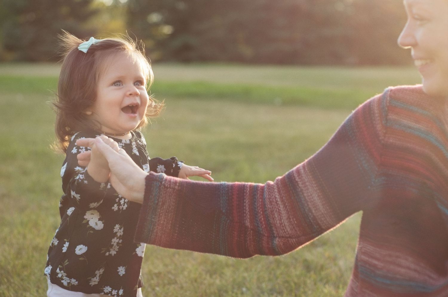 Baby holds her mother's hand as they both smile in a park.