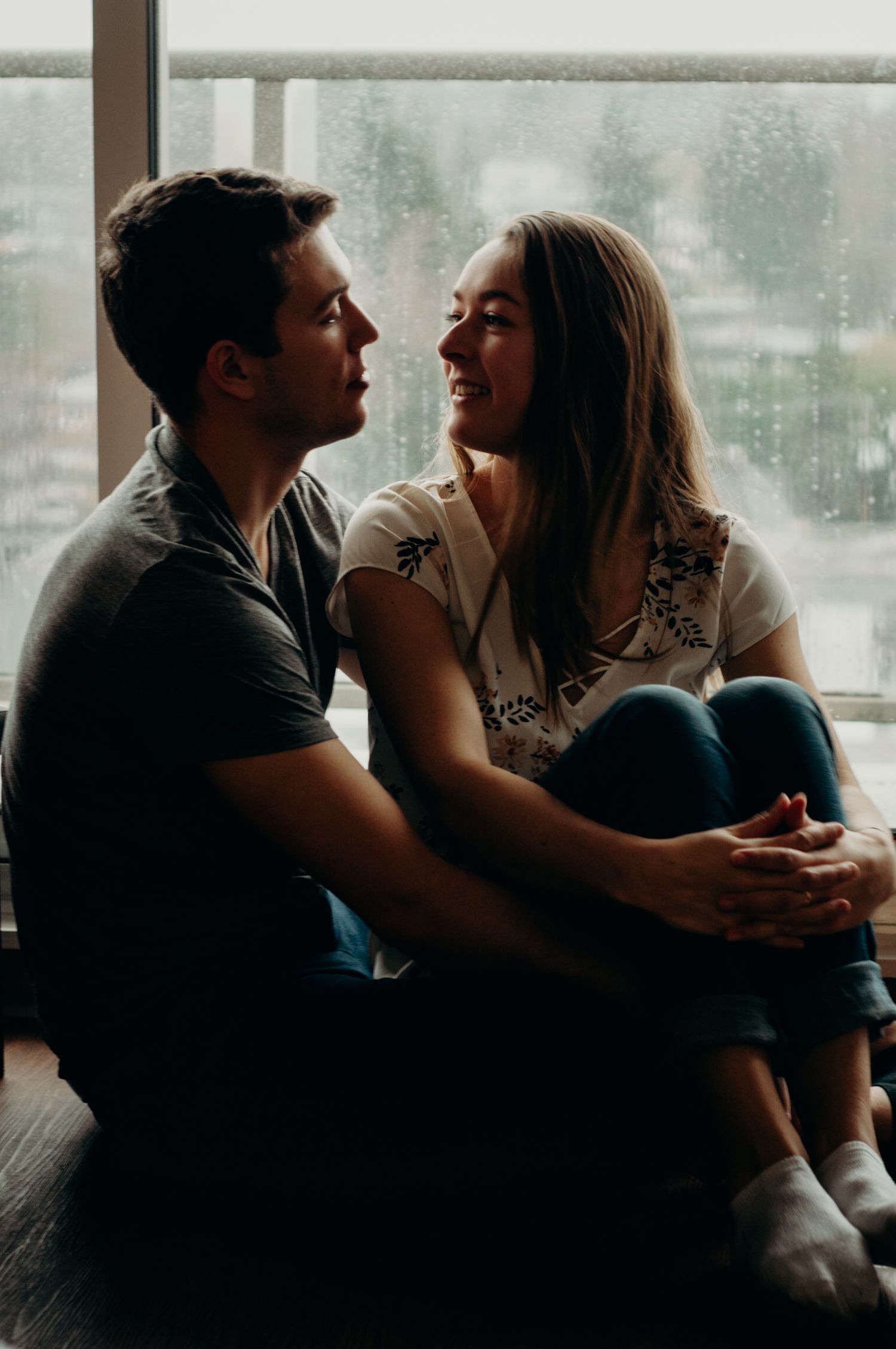 Couple looks at each other with love as they're cuddled up in front of a rainy window.