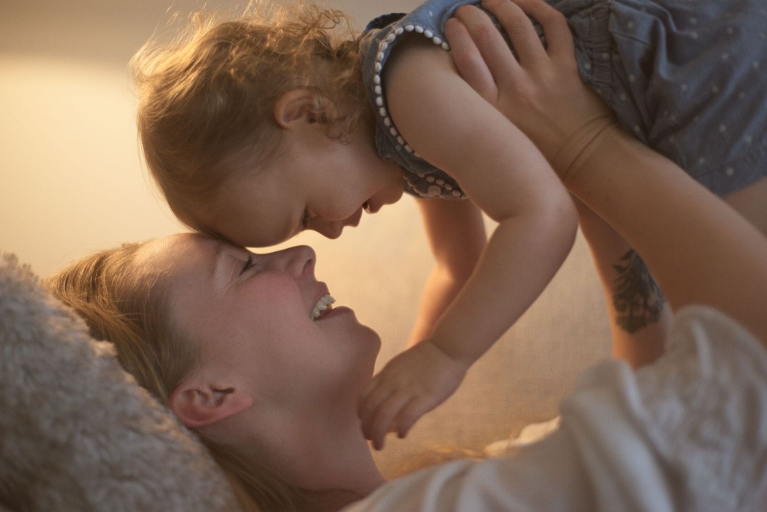 Mom and toddler laugh together as mom lifts her daughter up and they touch foreheads.