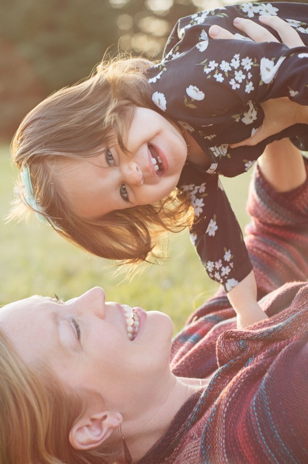 Baby and mom laugh as baby is lifted in to the air in a park.
