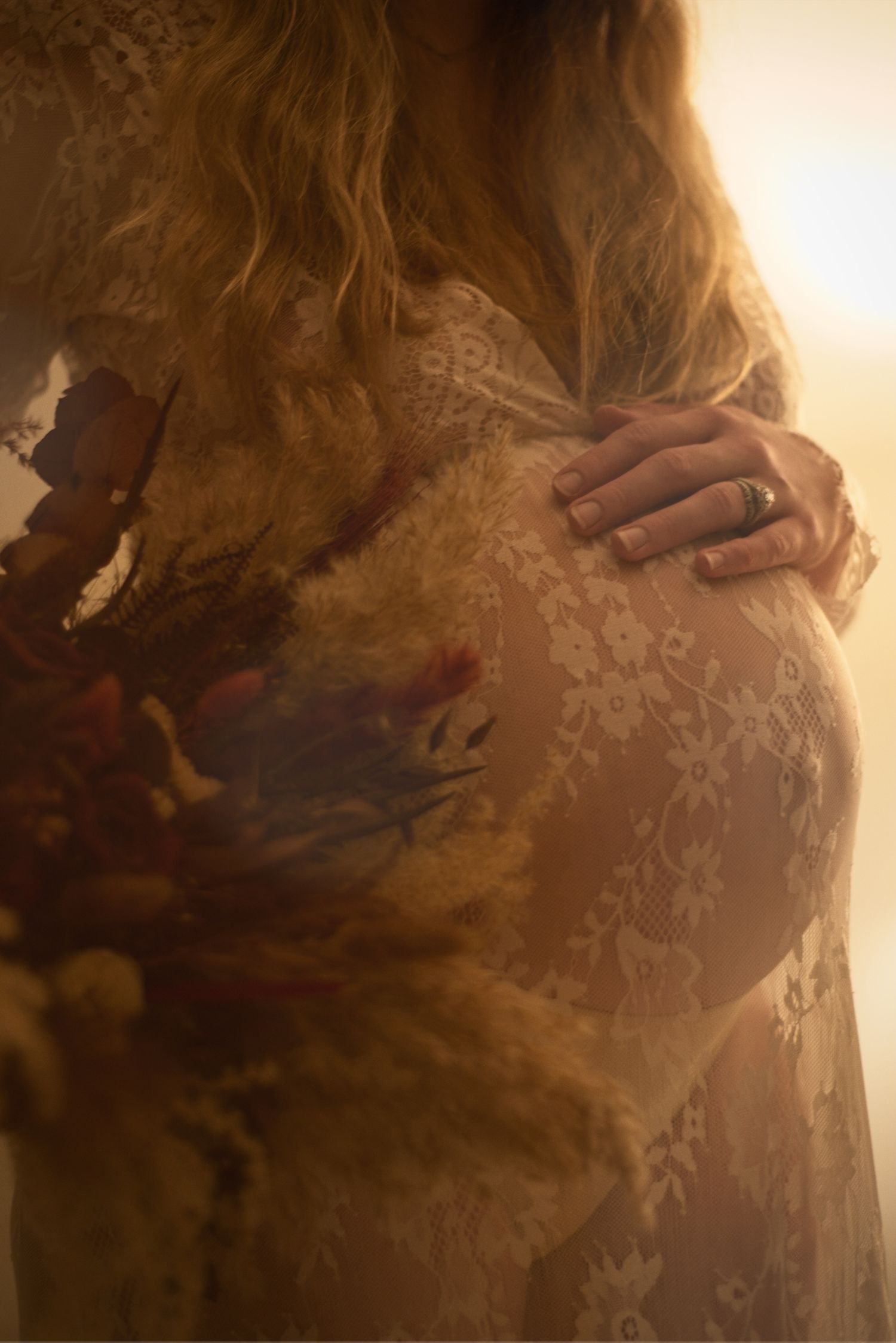 Pregnant woman rests her hand on her belly while wearing a lace cover and holding a wedding bouquet of flowers.