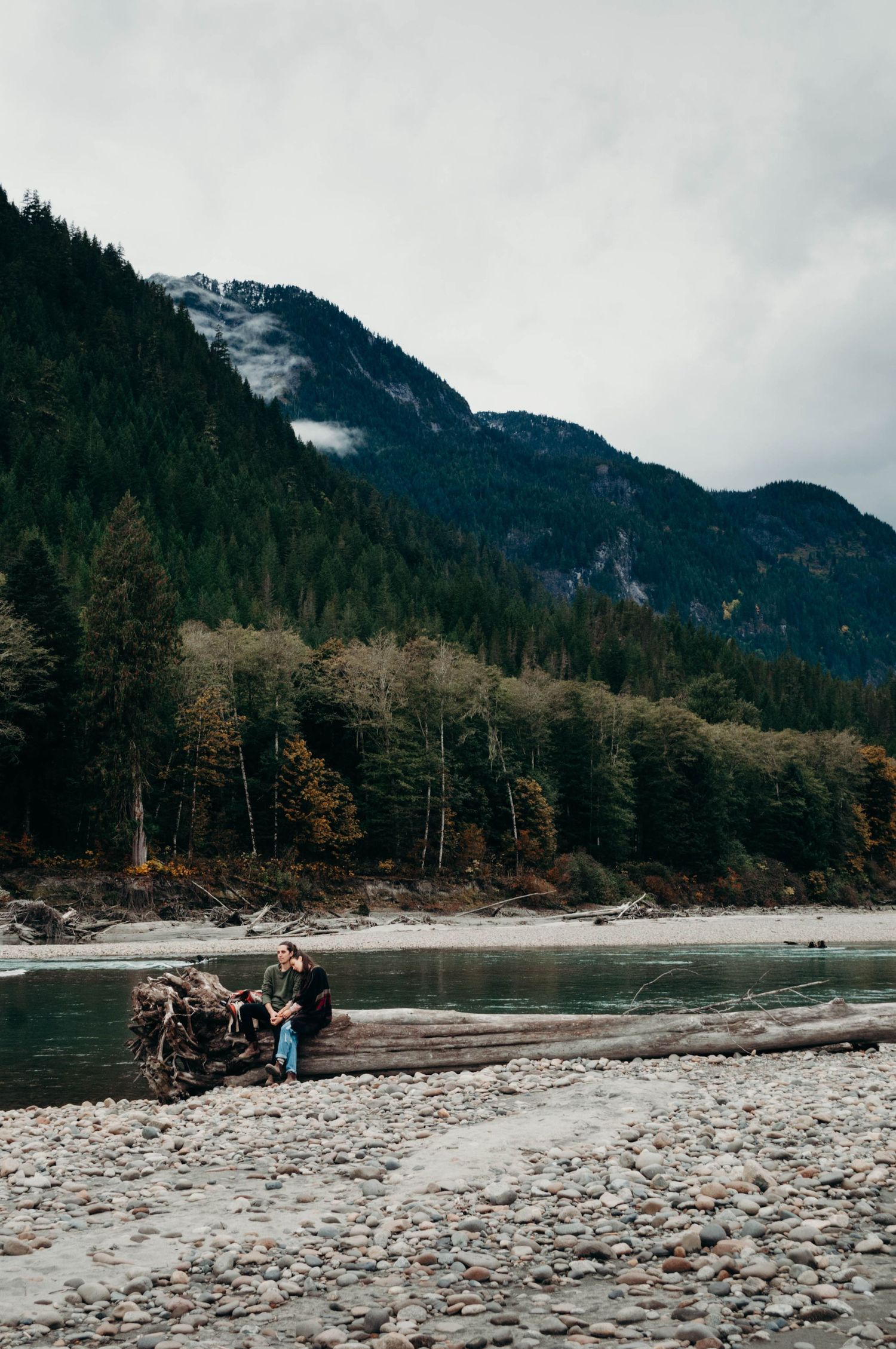 Engaged couple sit together on large log by a river.