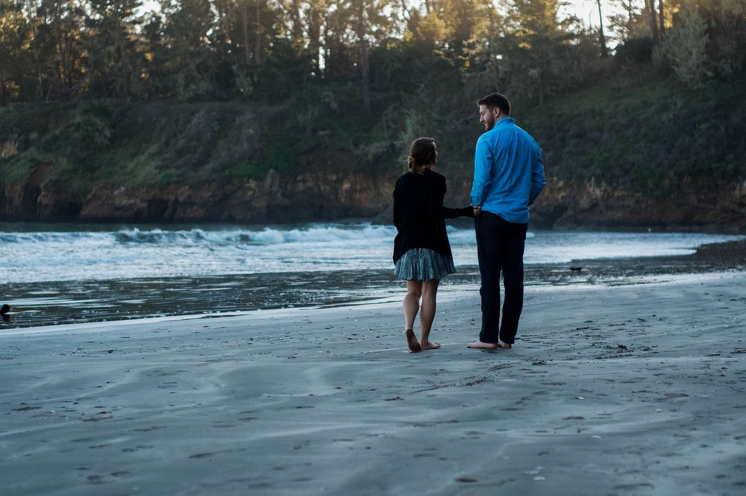 Newly engaged couple walks together barefoot along a beach during sunset.