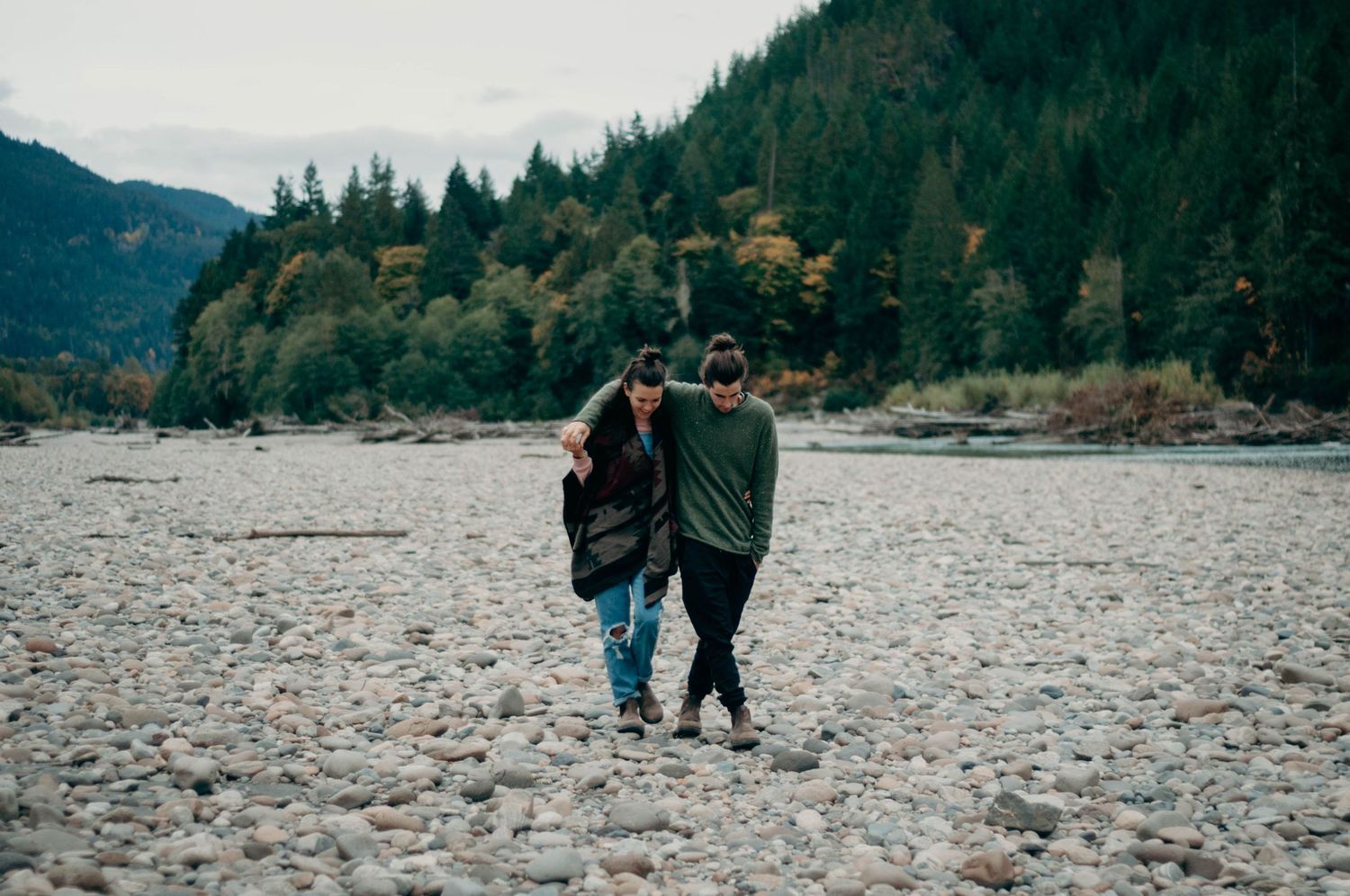 Fiance puts his arm around his partner as they walk along the rocks with the forest in the background.