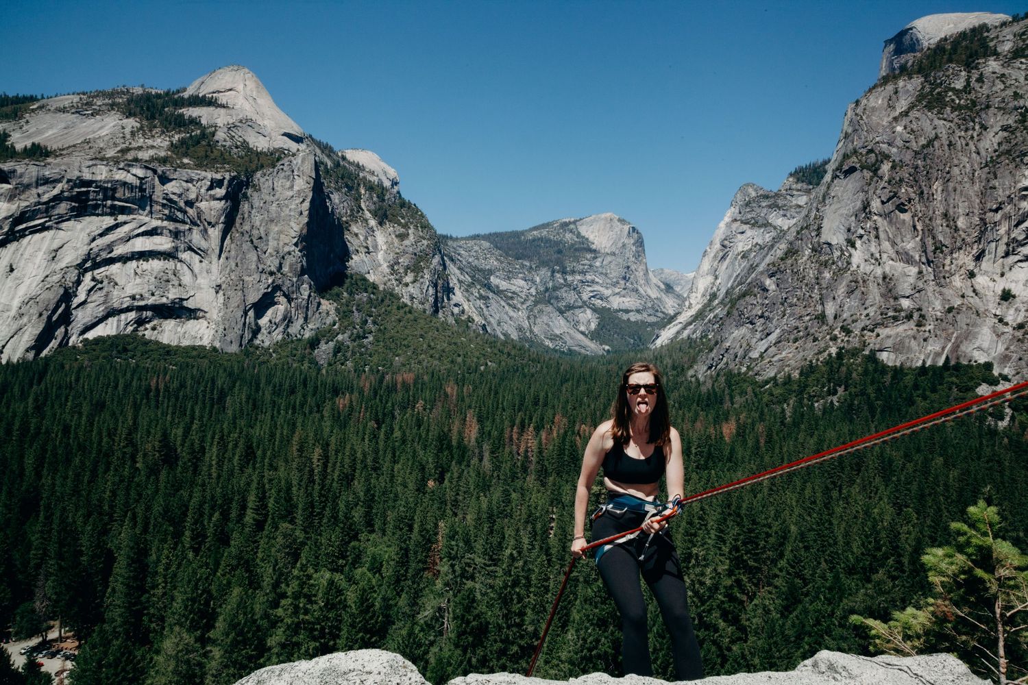 Photographer makes funny face at camera while rappelling down a rock in Yosemite National Park, California, with forest and mountains.