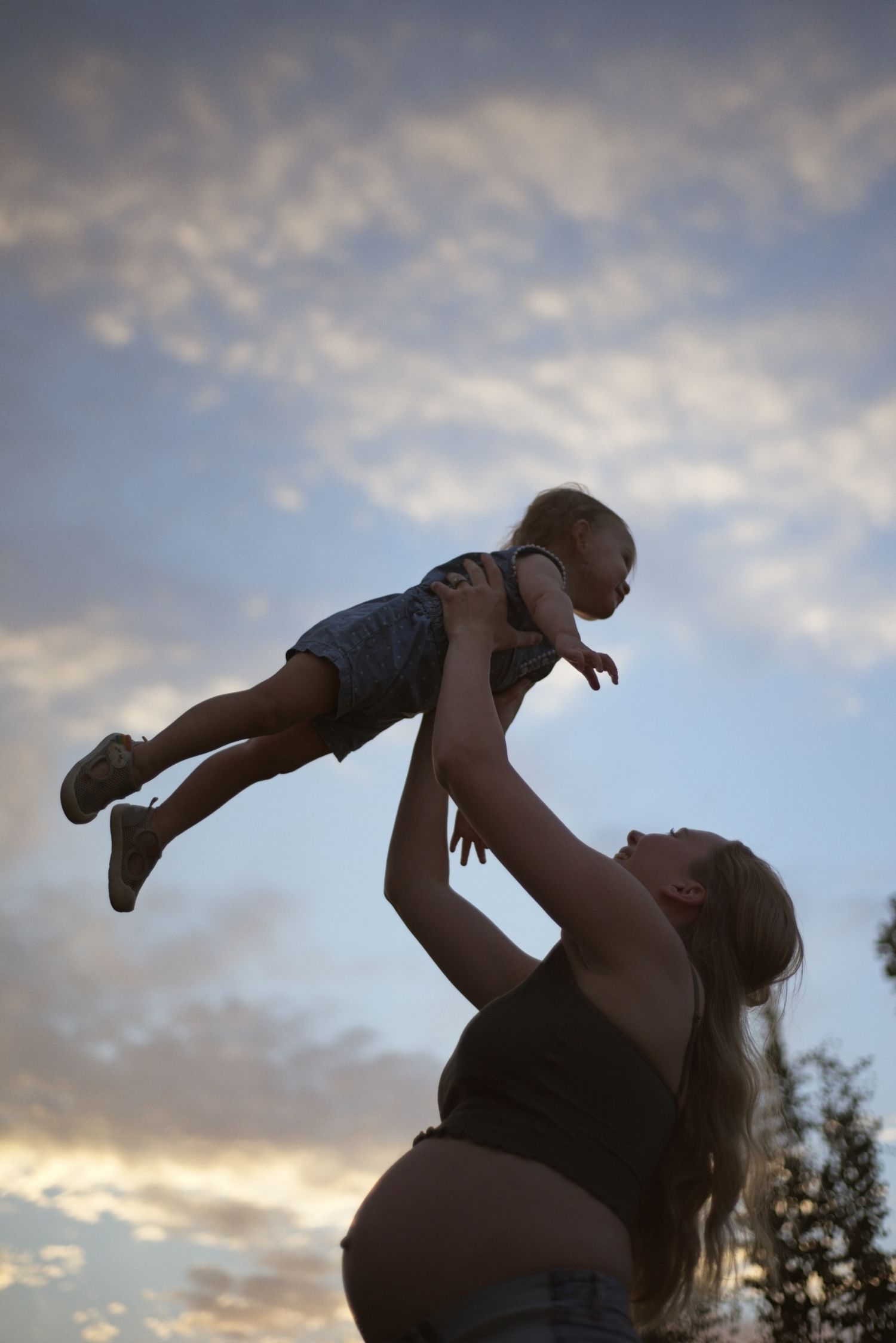 Pregnant mom lifts her toddler in to the air, silhouetted by the sunset sky.