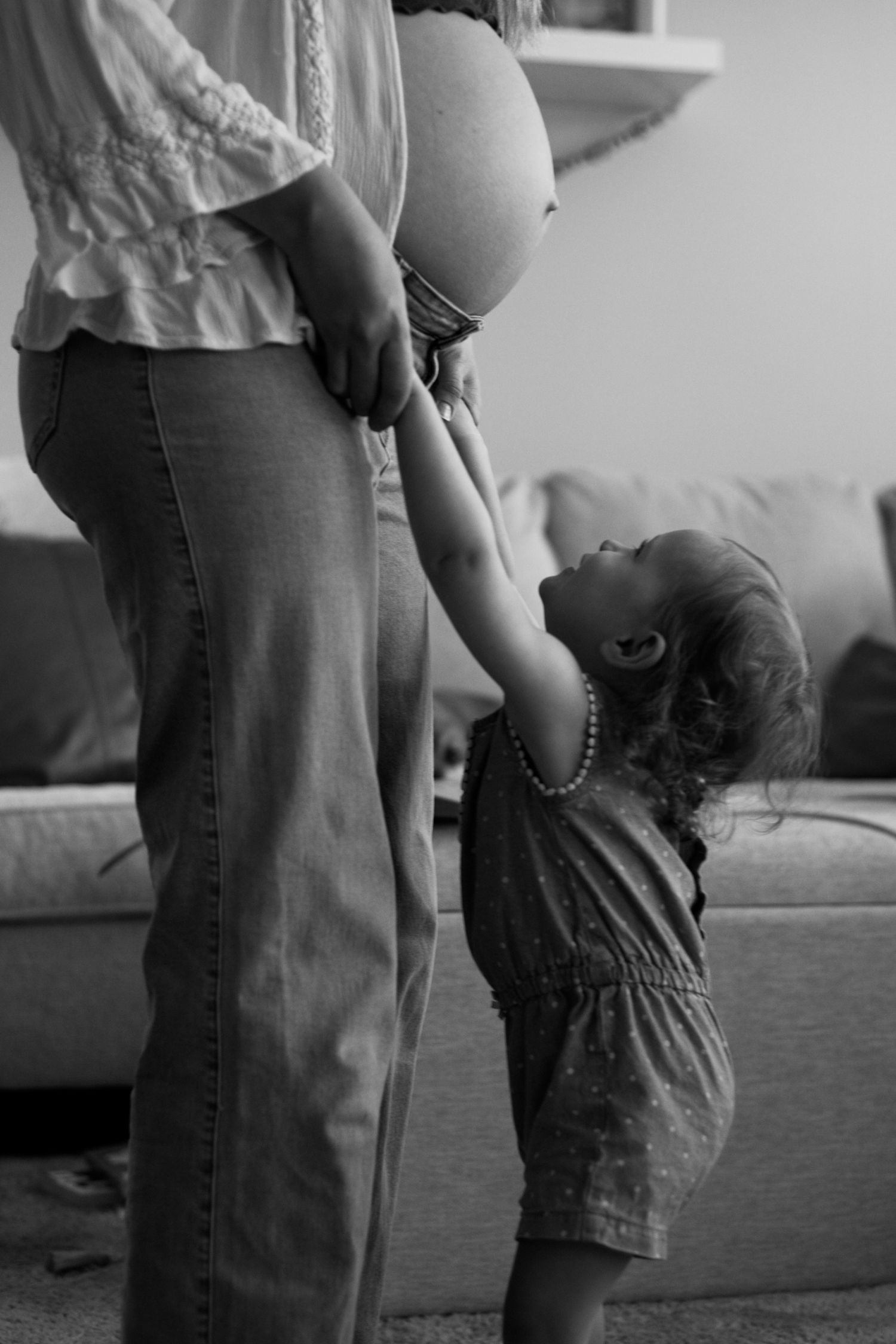 Pregnant mom holds the hands of her toddler while the toddler looks at her belly.