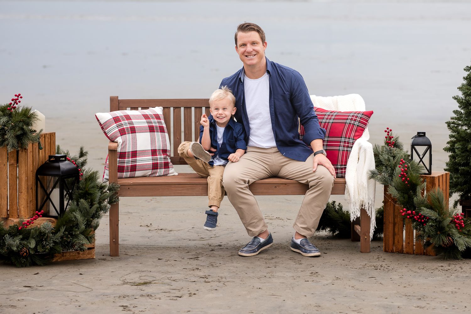 Family Christmas photo on beach with dad and toddler son on decorated bench with plaid pillows and holiday greenery.