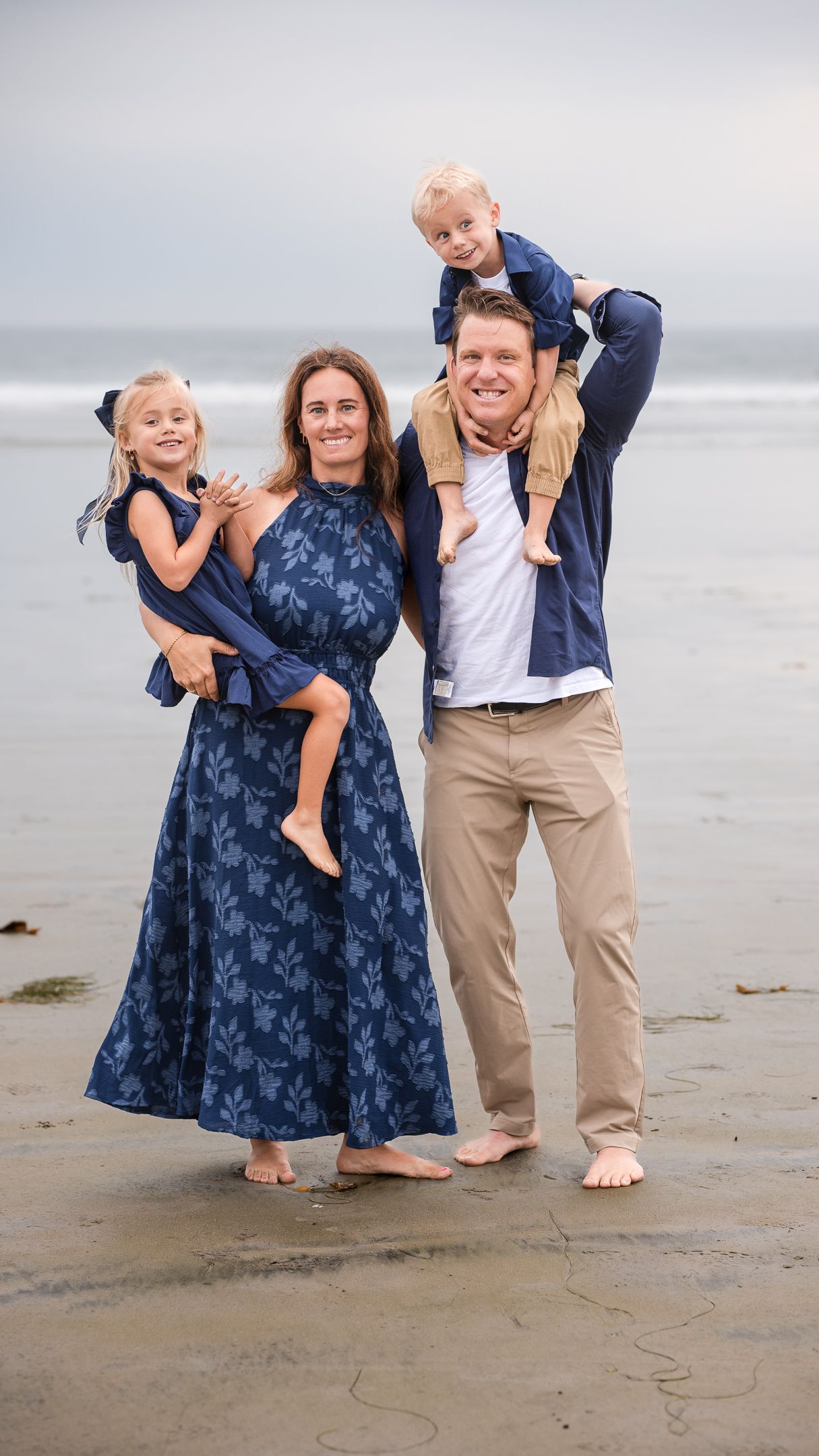 Happy family on beach with mother holding daughter and father carrying son on shoulders, all smiling.