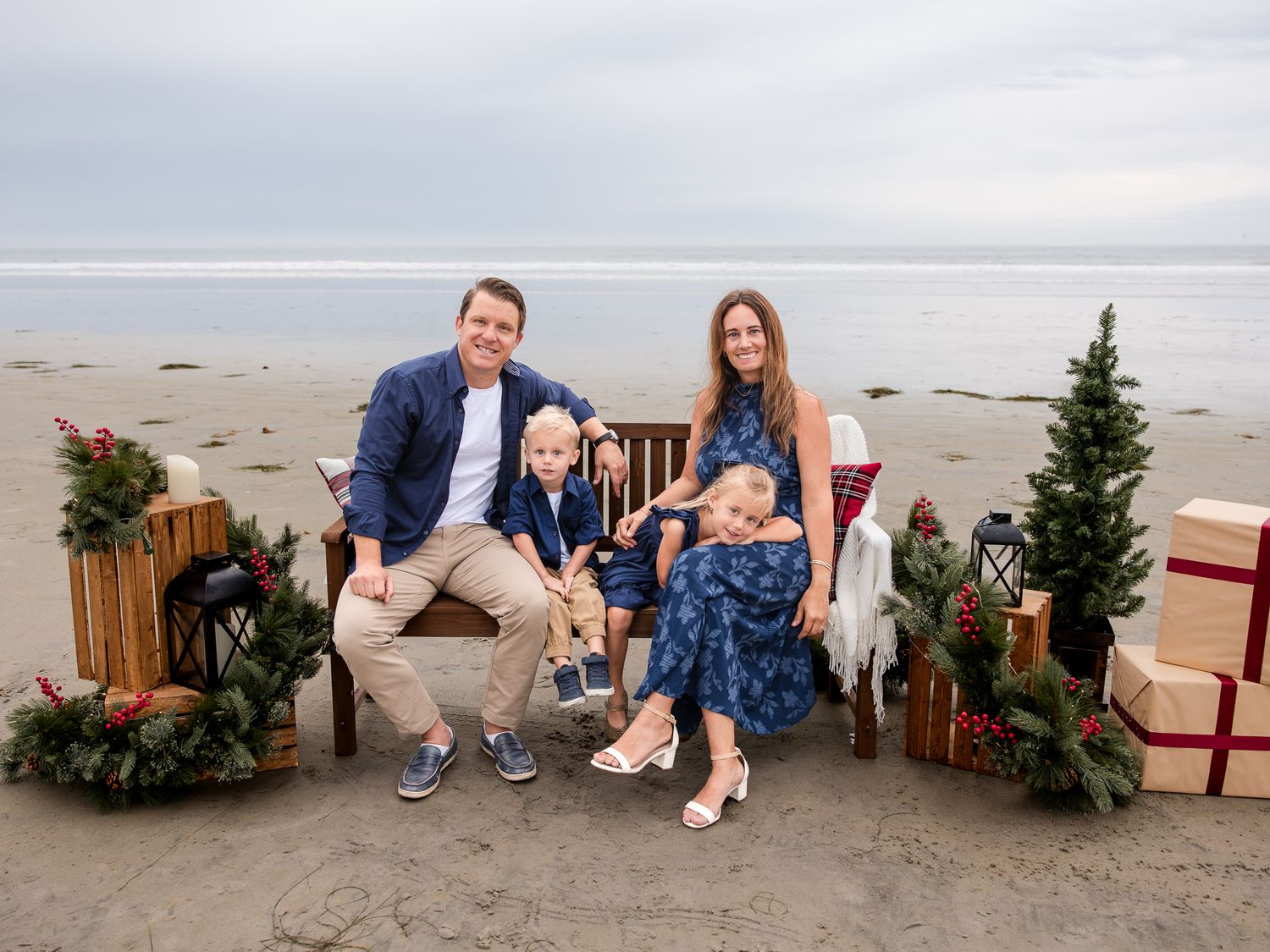 Family of four Christmas beach photo with holiday decorations, gifts, and mini Christmas trees on sand.