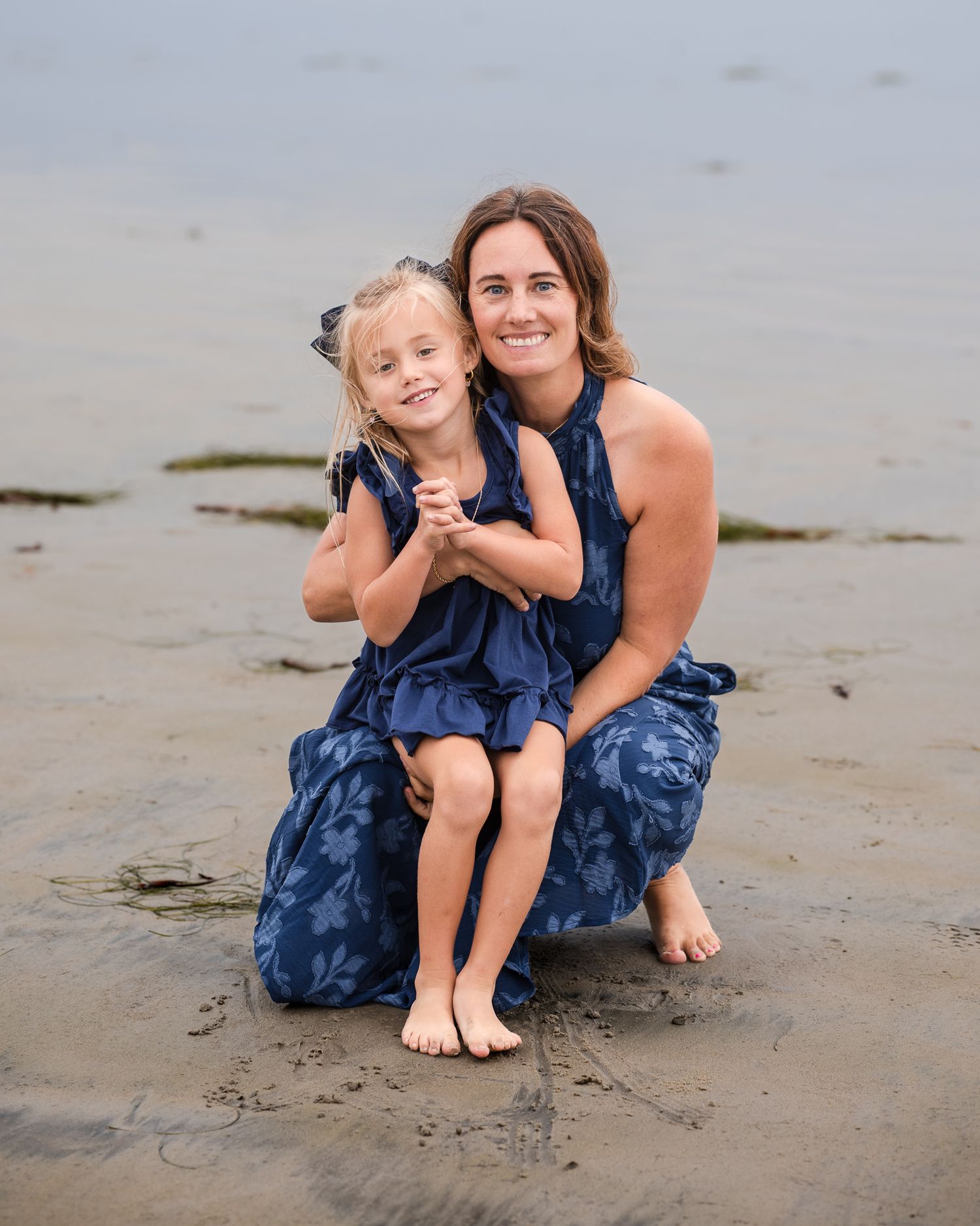 Mother and young blonde daughter in matching navy floral dresses posing together on sandy beach.