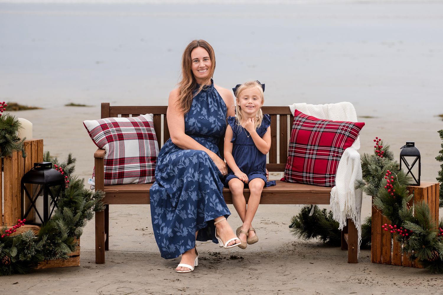 Mother and young daughter in matching navy floral dresses sit together on a Christmas-decorated beach bench.