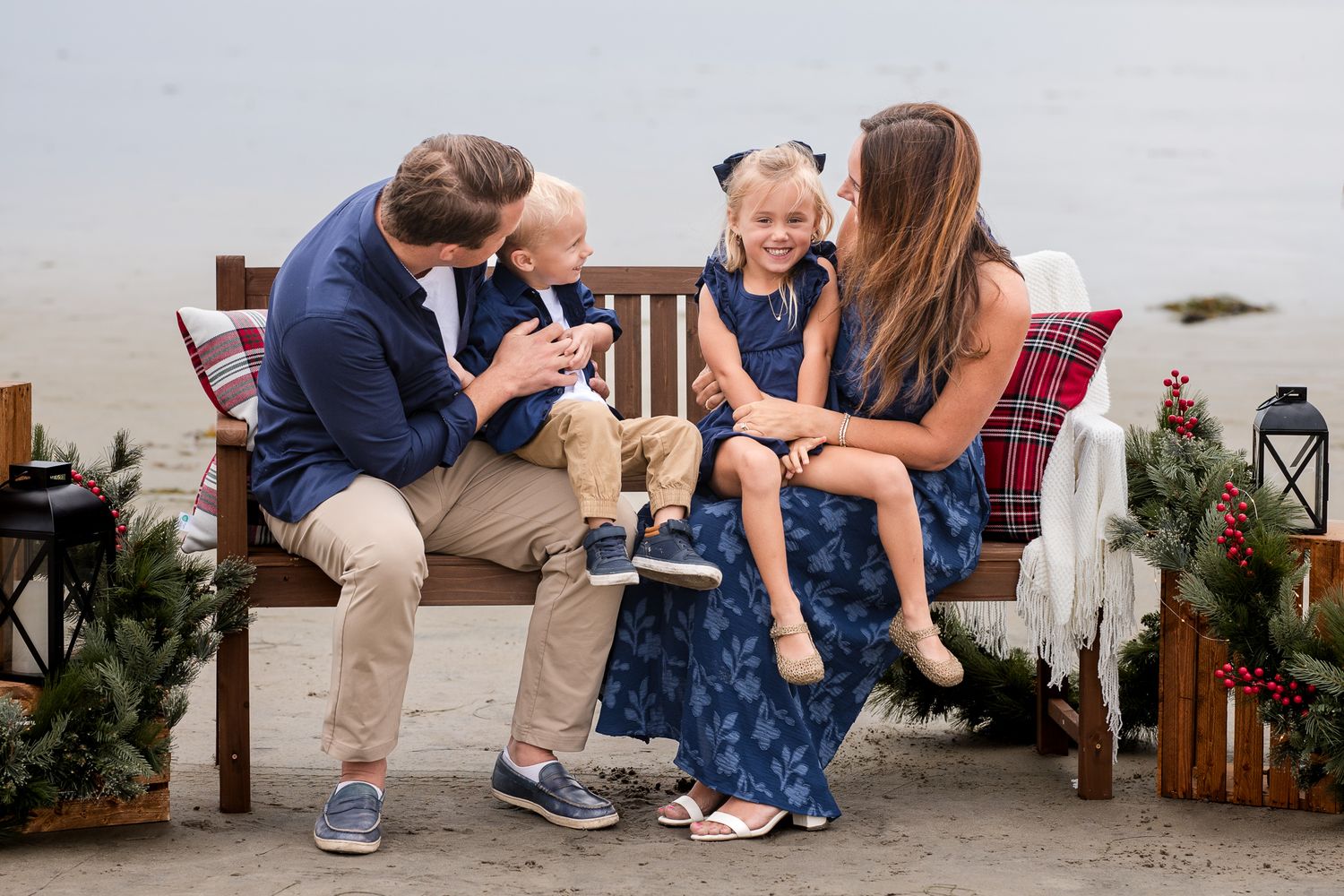 Family of four laughs together on a decorated Christmas bench on the beach in coordinated navy outfits.