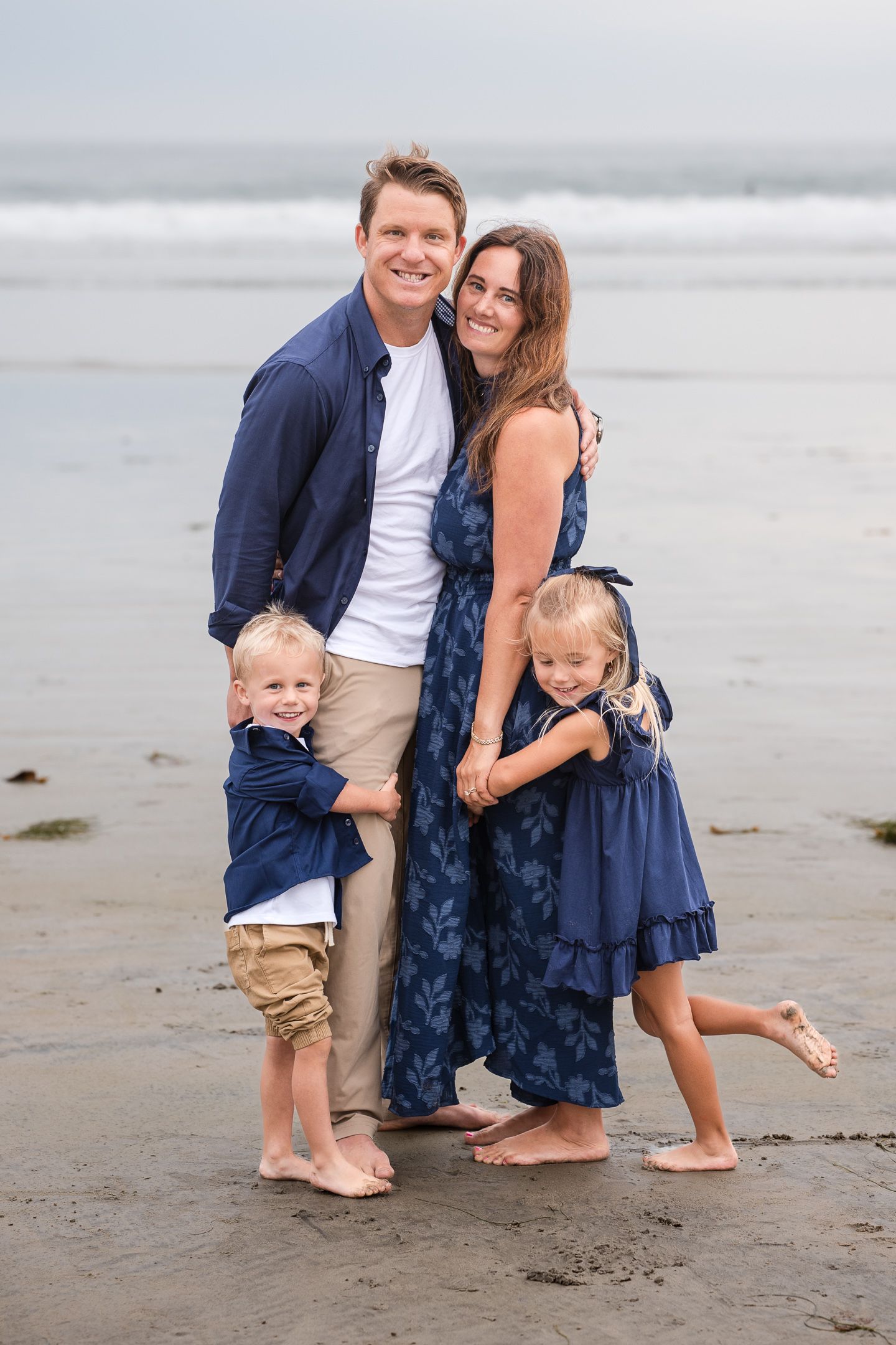 Family of four hugging on sandy beach, parents in navy outfits with two young blonde children smiling.