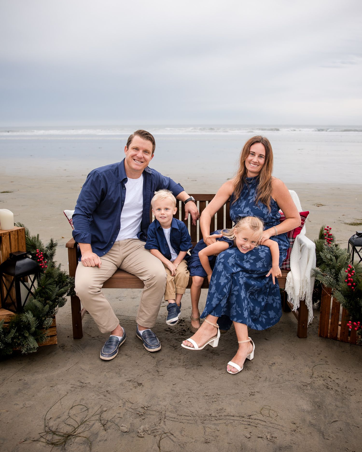 Family Christmas portrait on decorated beach bench with parents and two young children in navy outfits.