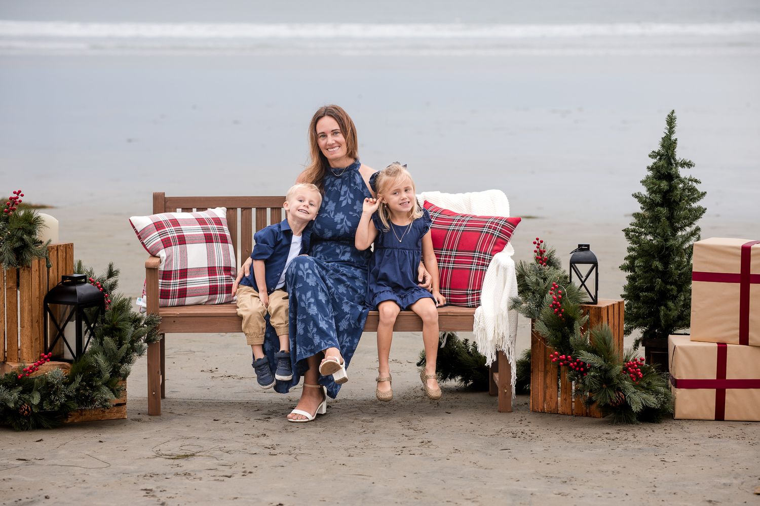 Mother sits with two young children on a Christmas-decorated beach bench, all wearing coordinated navy outfits.