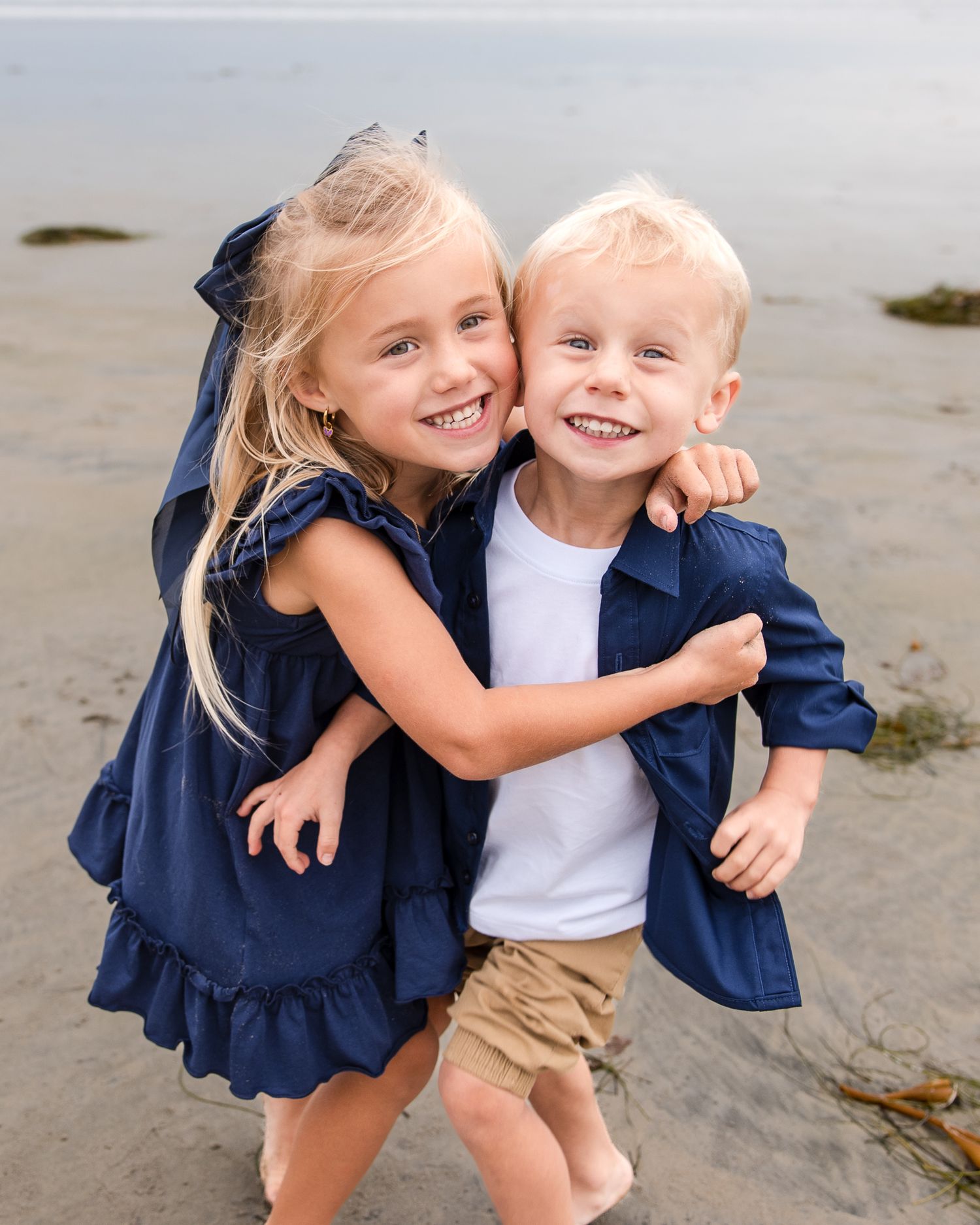 Two young blonde siblings in navy outfits hugging and smiling together on a sandy beach.