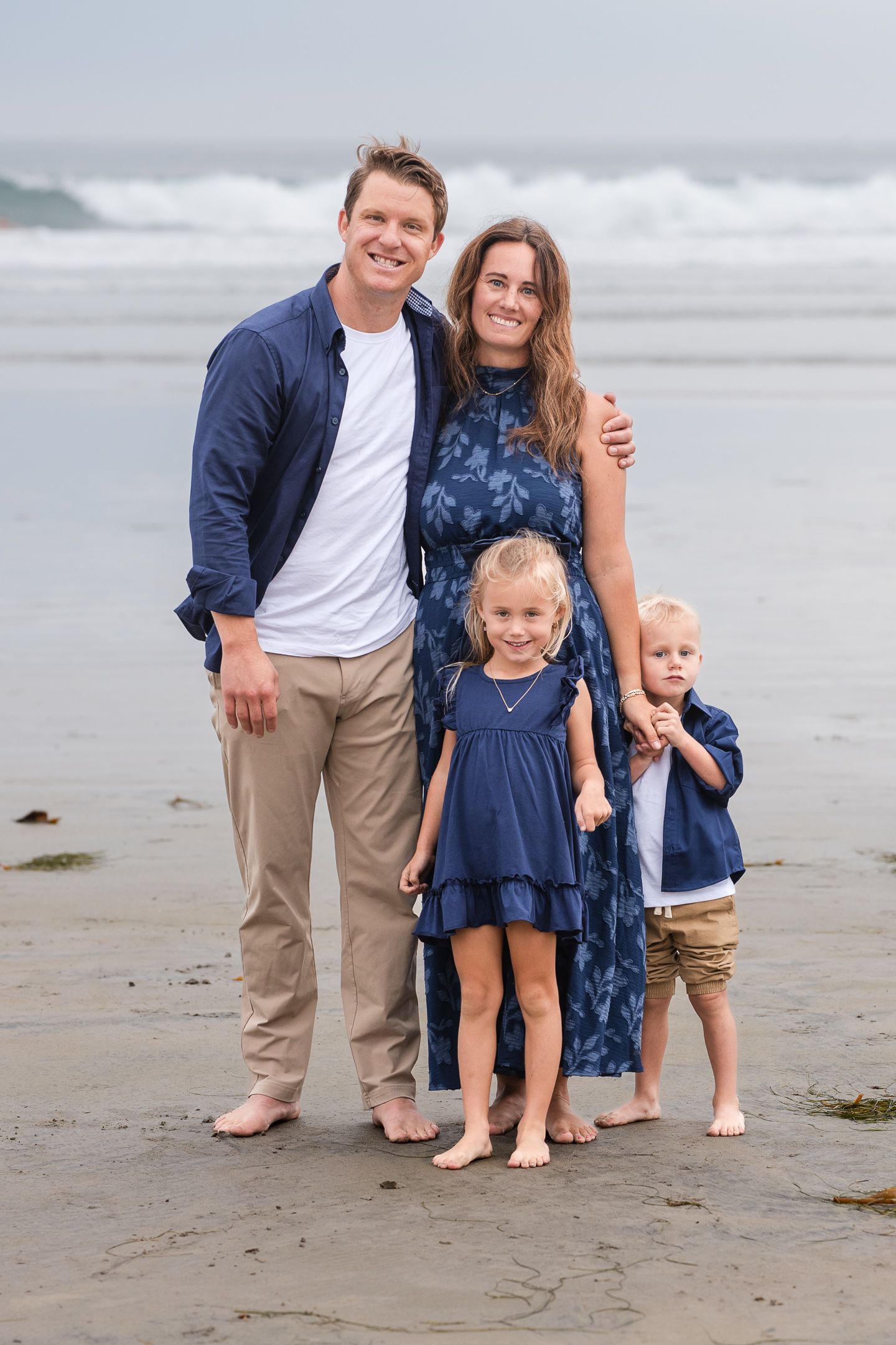 Family of four posing on beach, parents in navy outfits with two young blonde children standing in front.