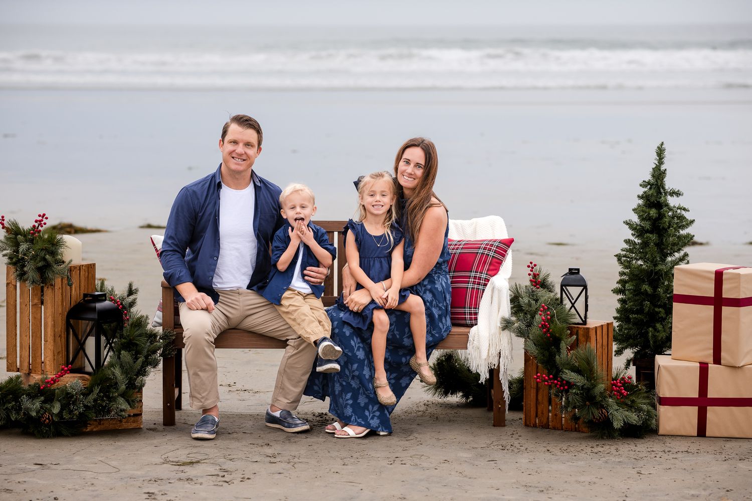 Family of four smiles together on a beach bench surrounded by holiday Christmas decorations and gifts.
