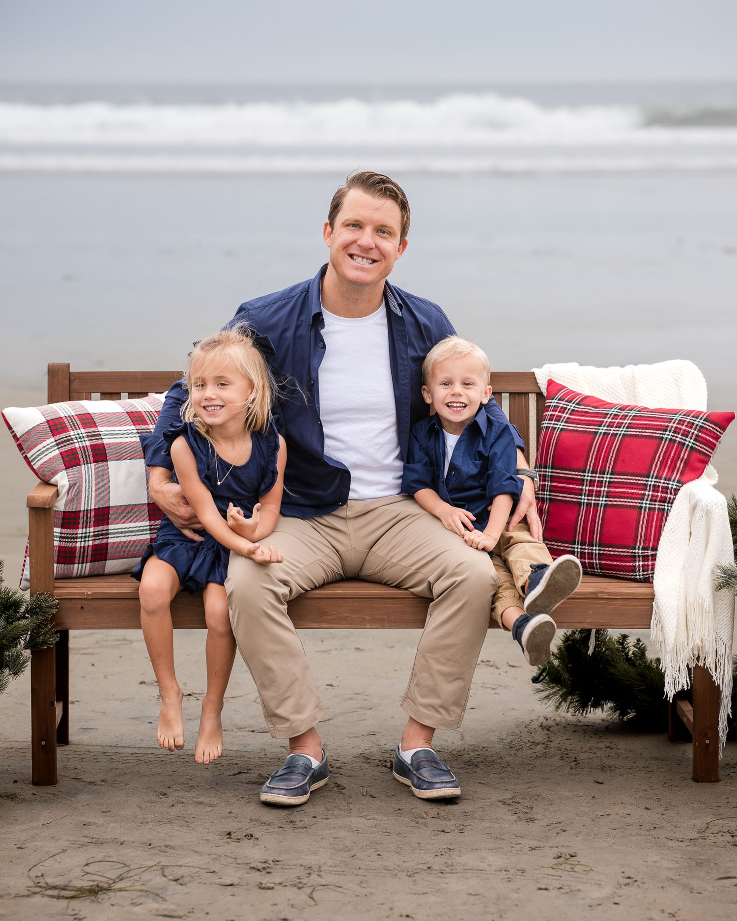 Father poses with two young children on a Christmas-decorated beach bench, all wearing coordinated navy outfits.