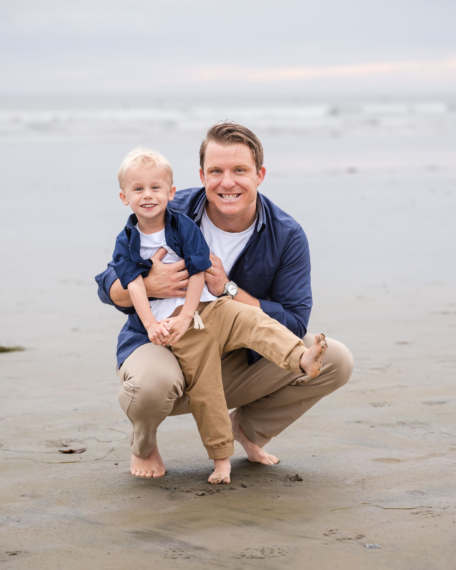 Father crouching on sandy beach holding young blonde toddler son, both smiling in navy and khaki outfits.