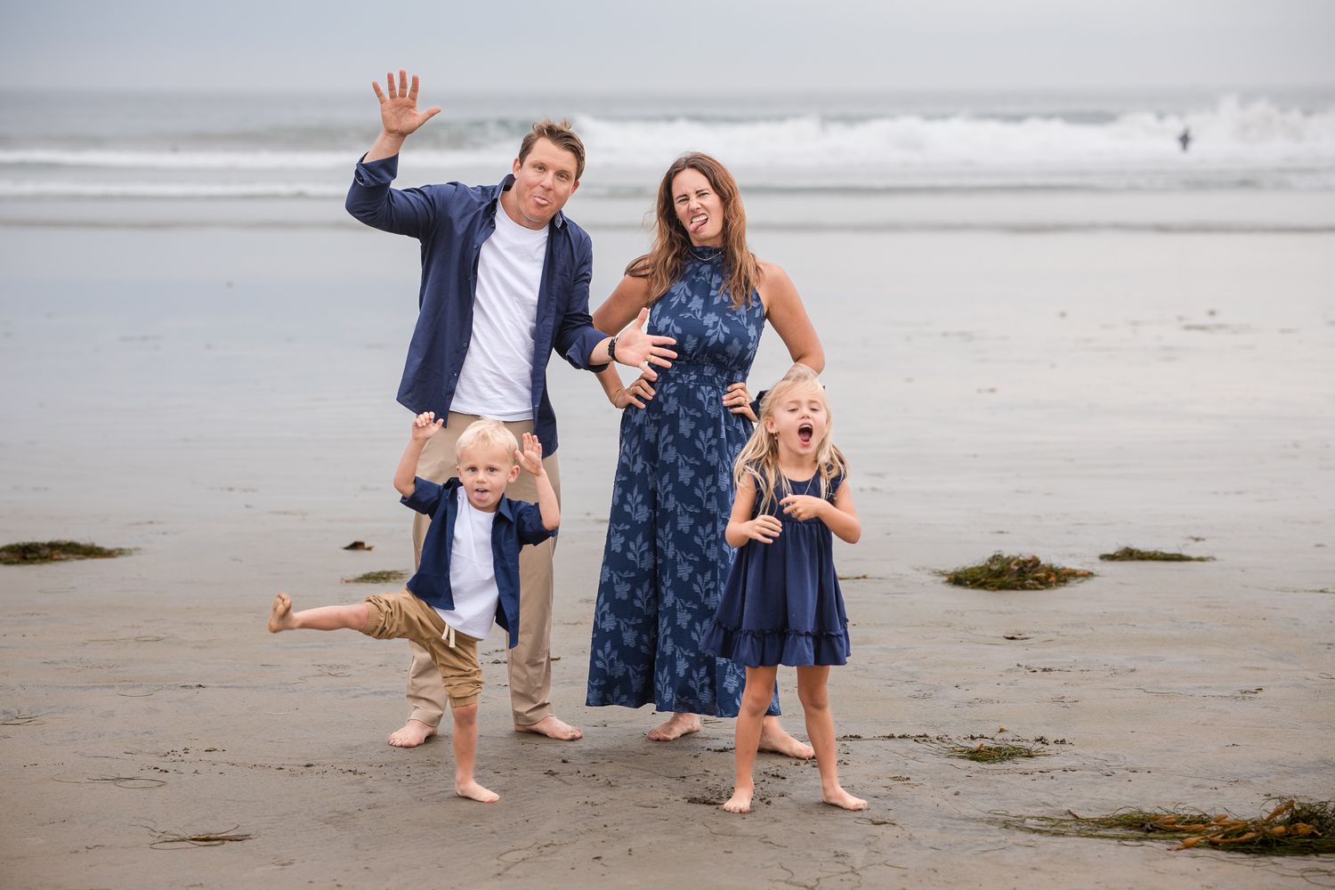 Playful family of four on beach with dad waving, kids jumping, and mom in navy floral dress laughing.