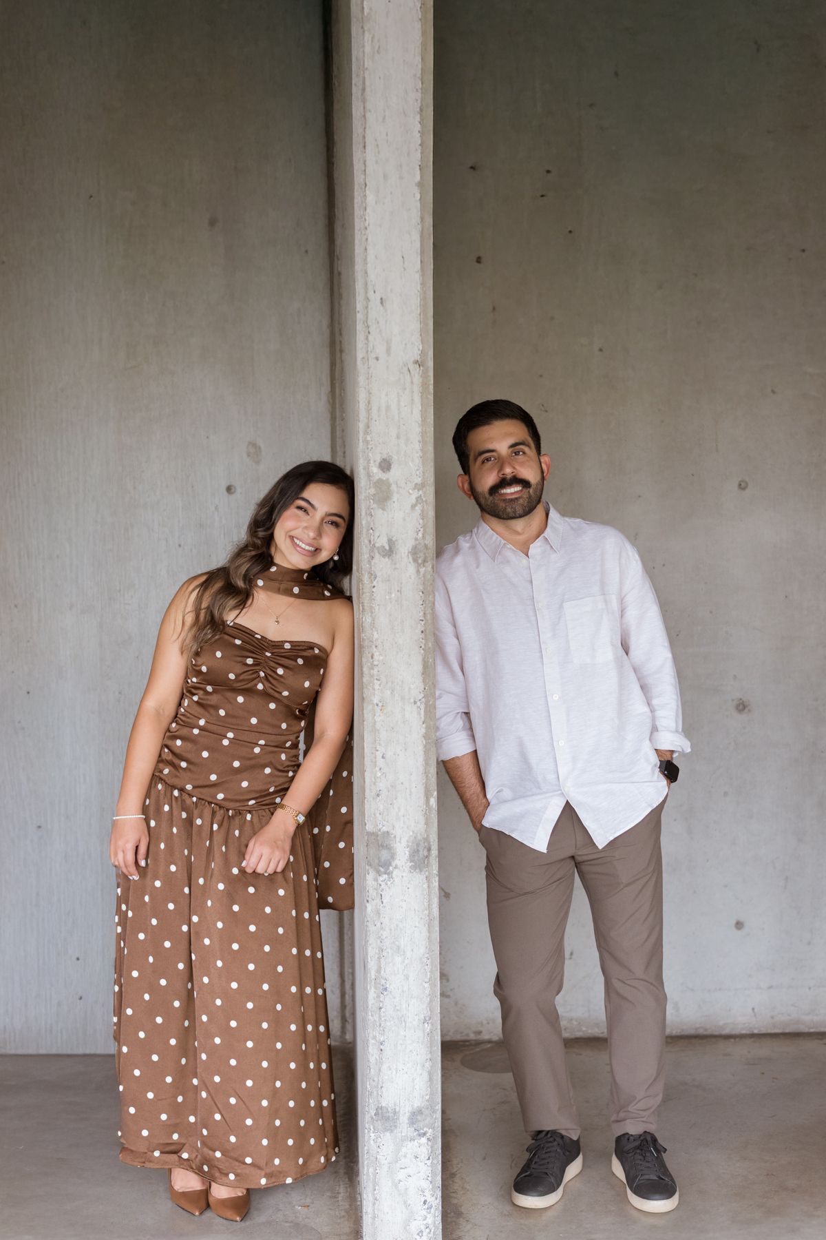 Couple posing by a concrete pillar, woman in brown polka dot dress, man in white linen shirt.
