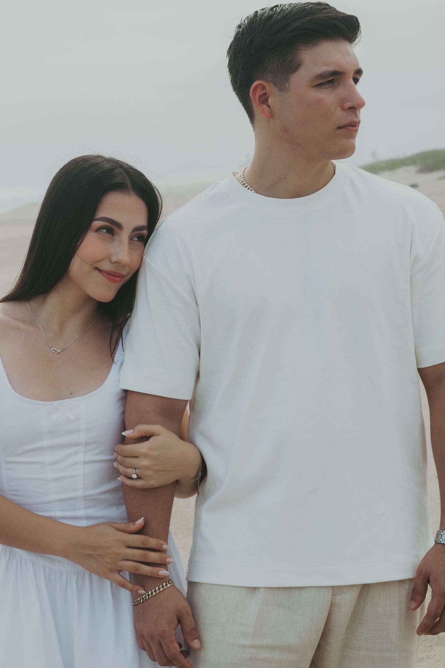 Couple in white outfits posing on a beach, woman in dress leaning on man in t-shirt and linen pants.
