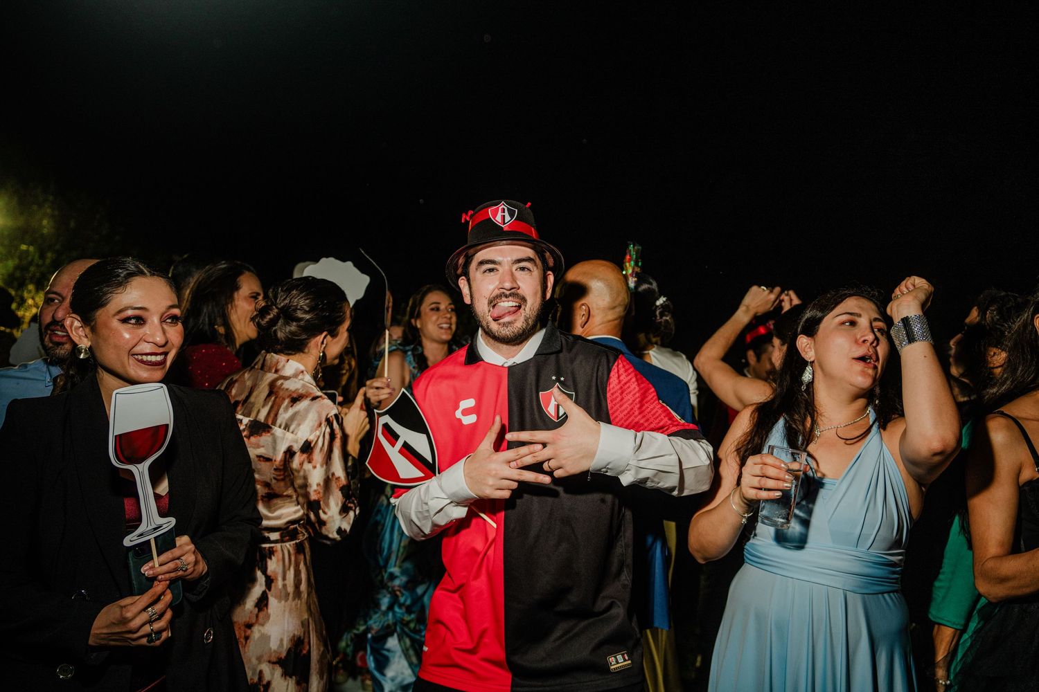 Man in soccer jersey dancing joyfully with guests at outdoor wedding reception party at night.