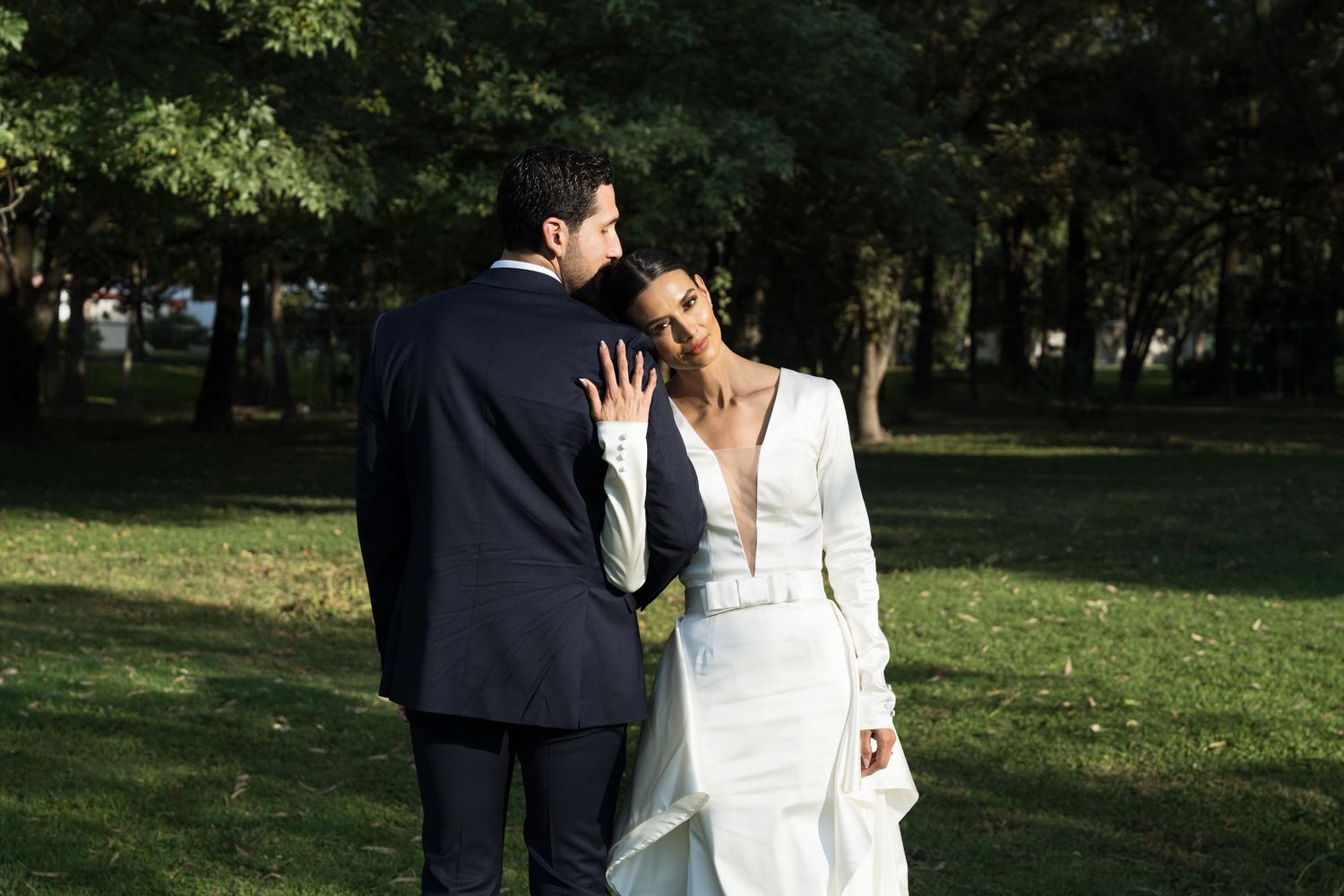 Bride in white long-sleeve gown smiling at groom while walking in sunny park.