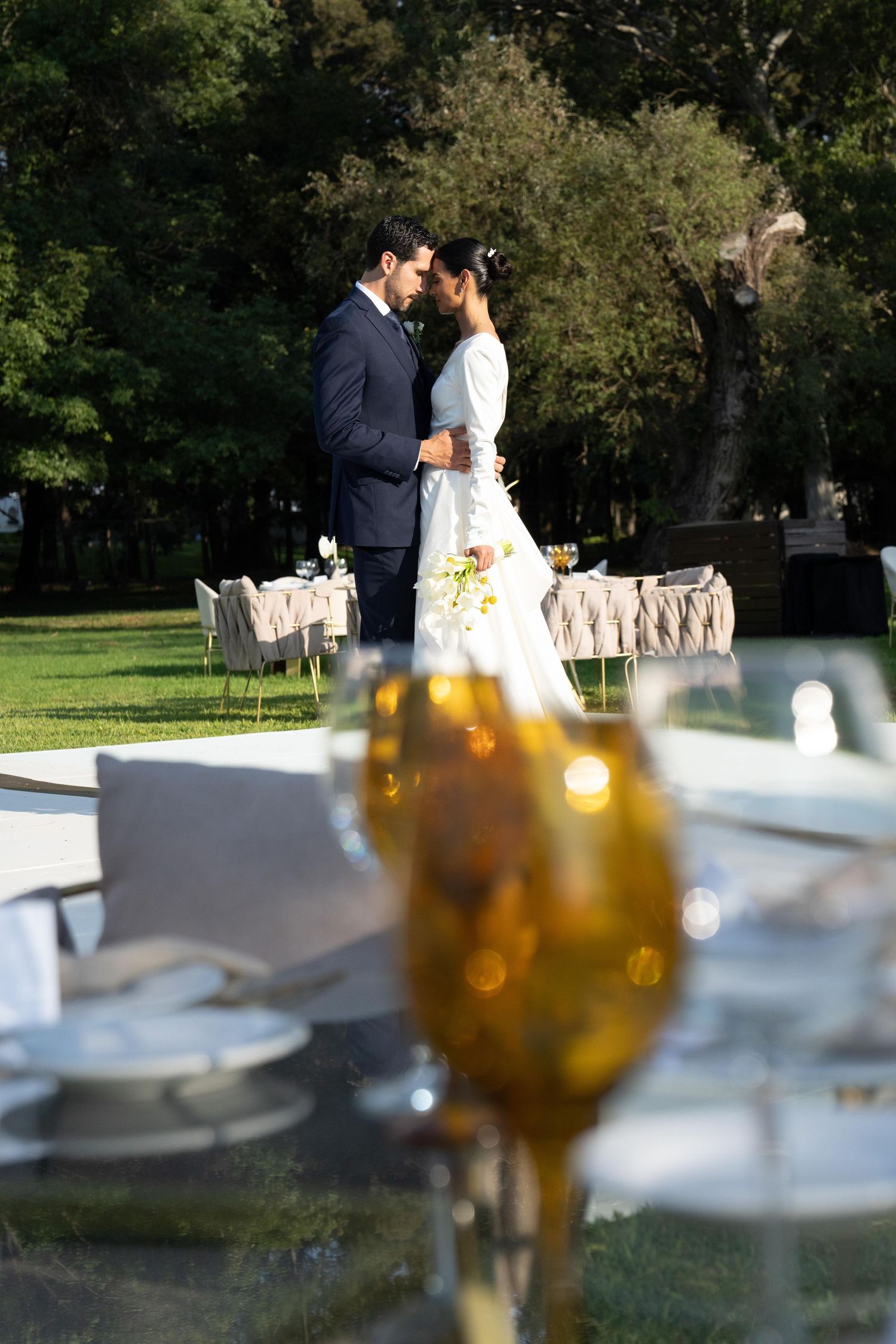 Newlyweds sharing intimate moment near outdoor reception tables with bokeh foreground.