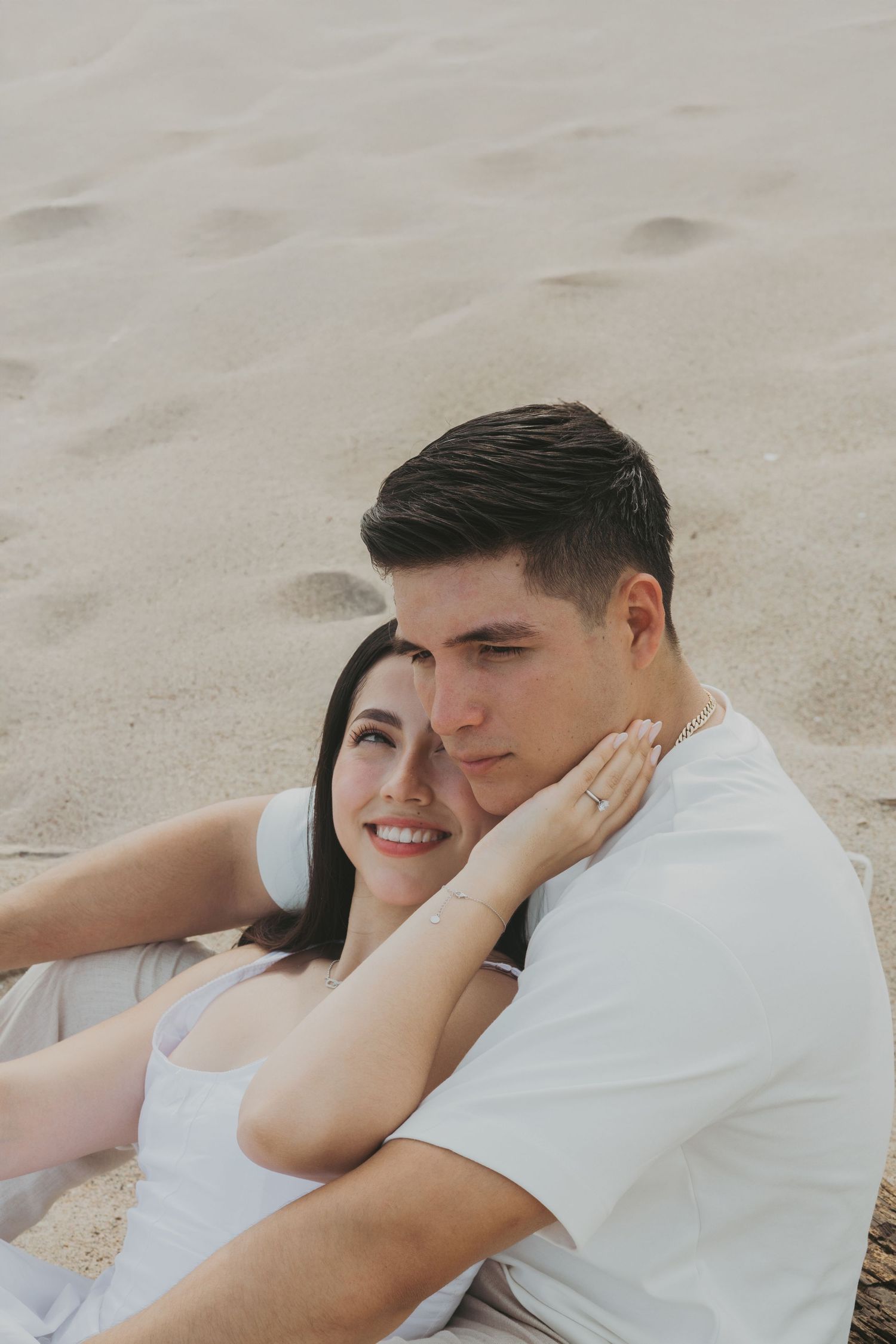 Couple sitting on sandy beach, woman smiling in white dress leaning against man in white shirt.